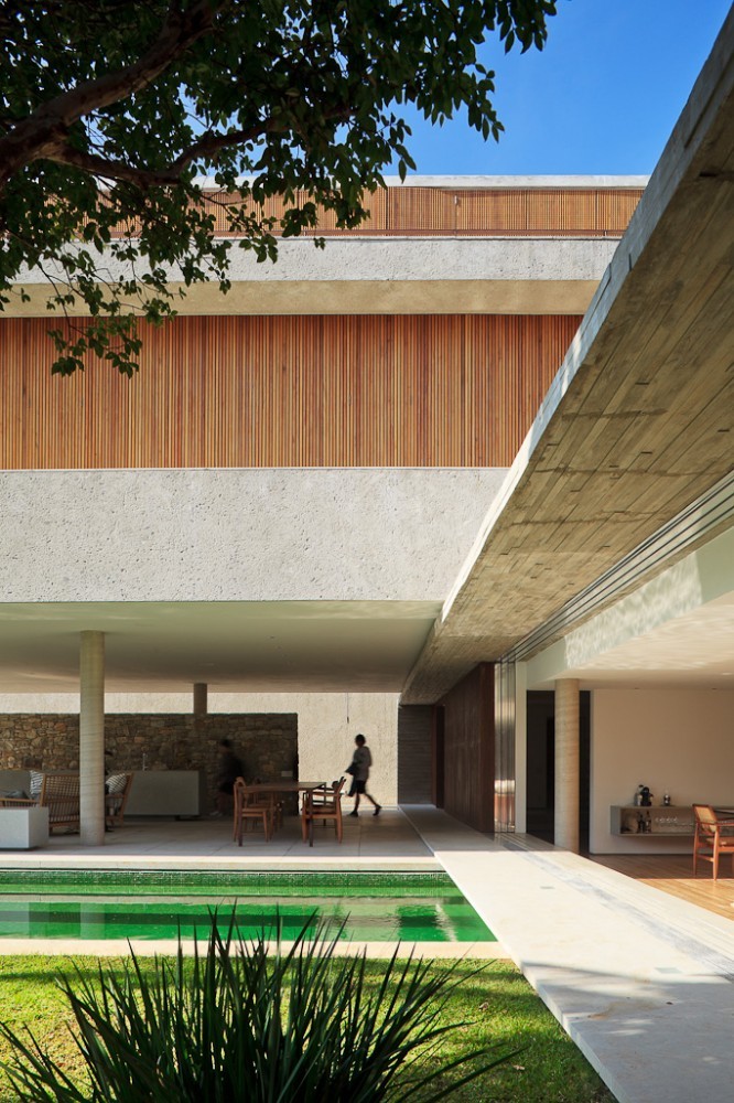 Concrete slab overhang and slatted timber upper volume above a covered terrace, with a person walking past a dining table beside the green pool