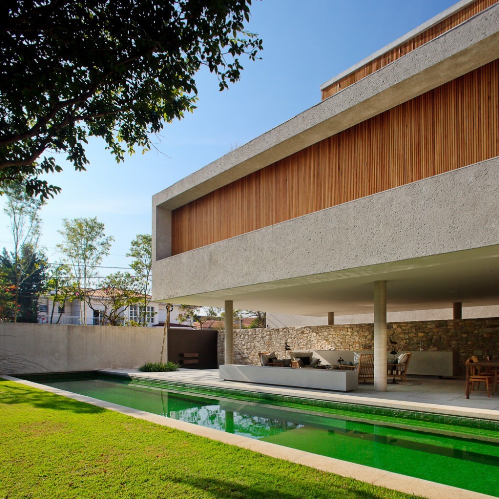 Cantilevered concrete volume with vertical timber slats rising above a stone wall and lounge area, seen across the lawn and green pool under blue sky