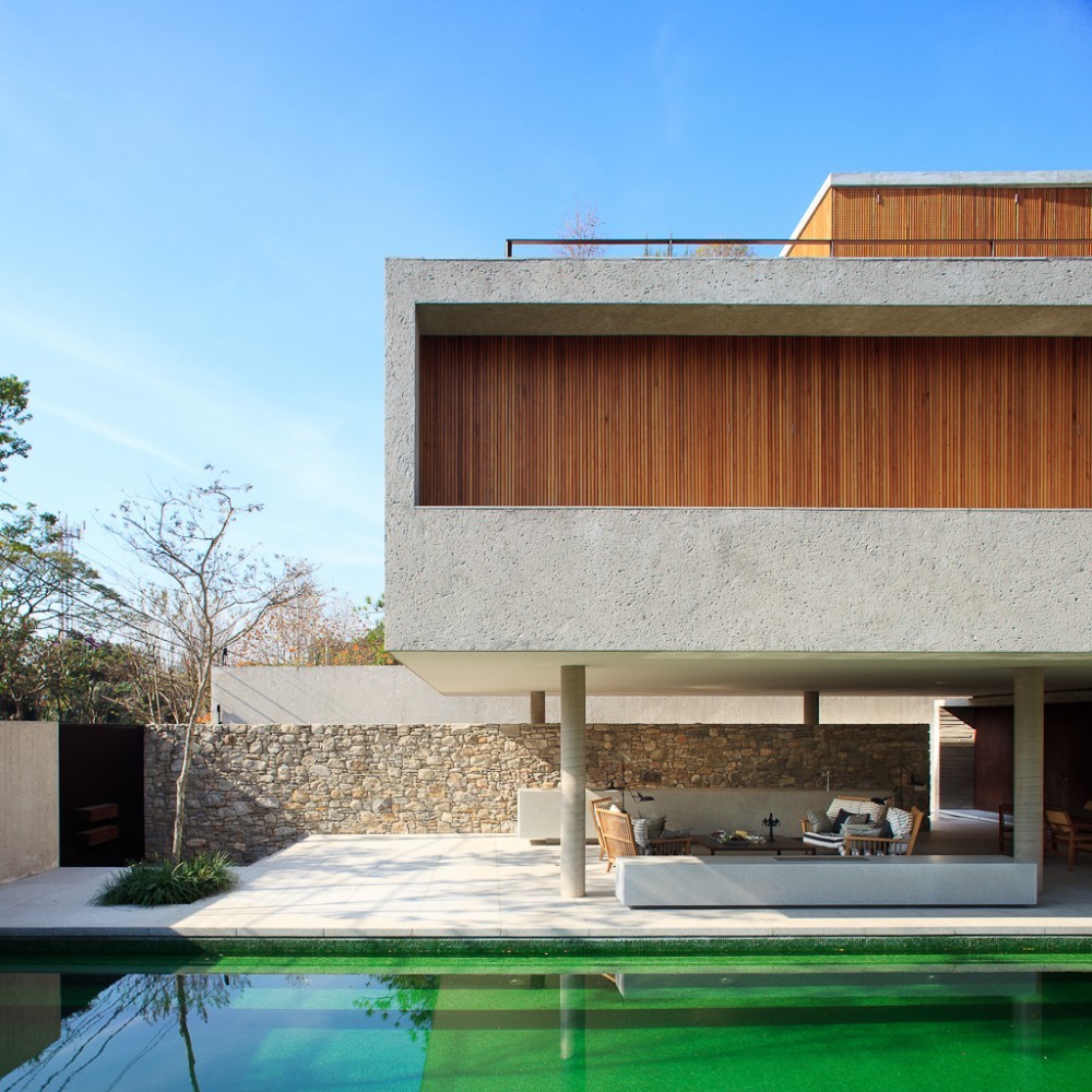 Frontal view of the raised concrete box with timber slat band, slender columns and a rubble stone wall behind the pool in clear daylight