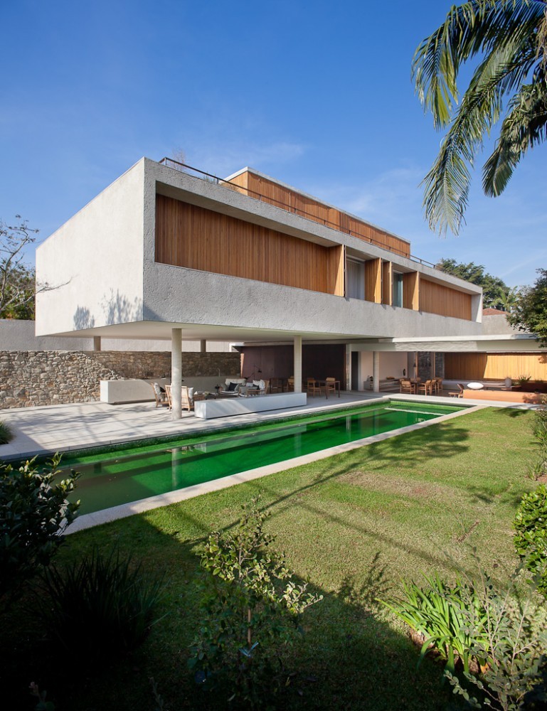 Corner view of the two-storey concrete and timber house above the long green pool, with a palm frond in the upper corner