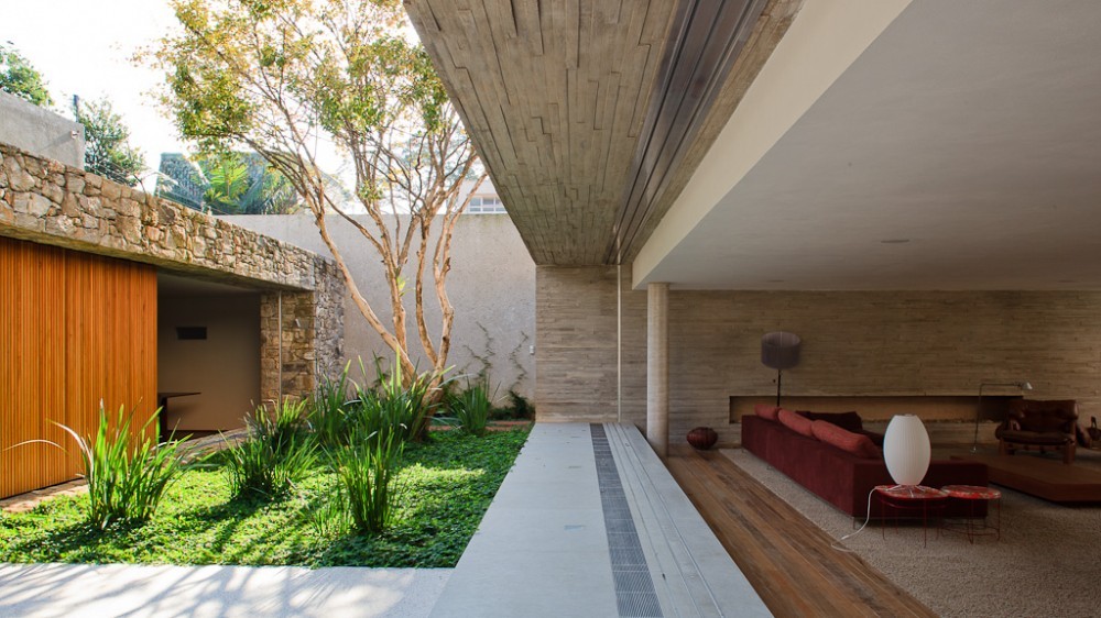 Planted courtyard with grasses and a slender tree beside a stone wall, opening to a living room with red sofa beneath a board-marked concrete soffit