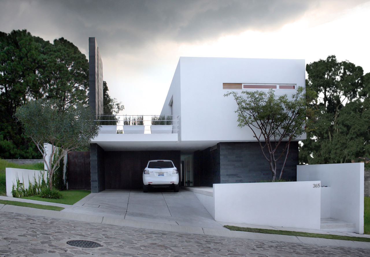 Street facade of a white cantilevered upper volume over a dark stone carport with a white car, under a stormy grey sky