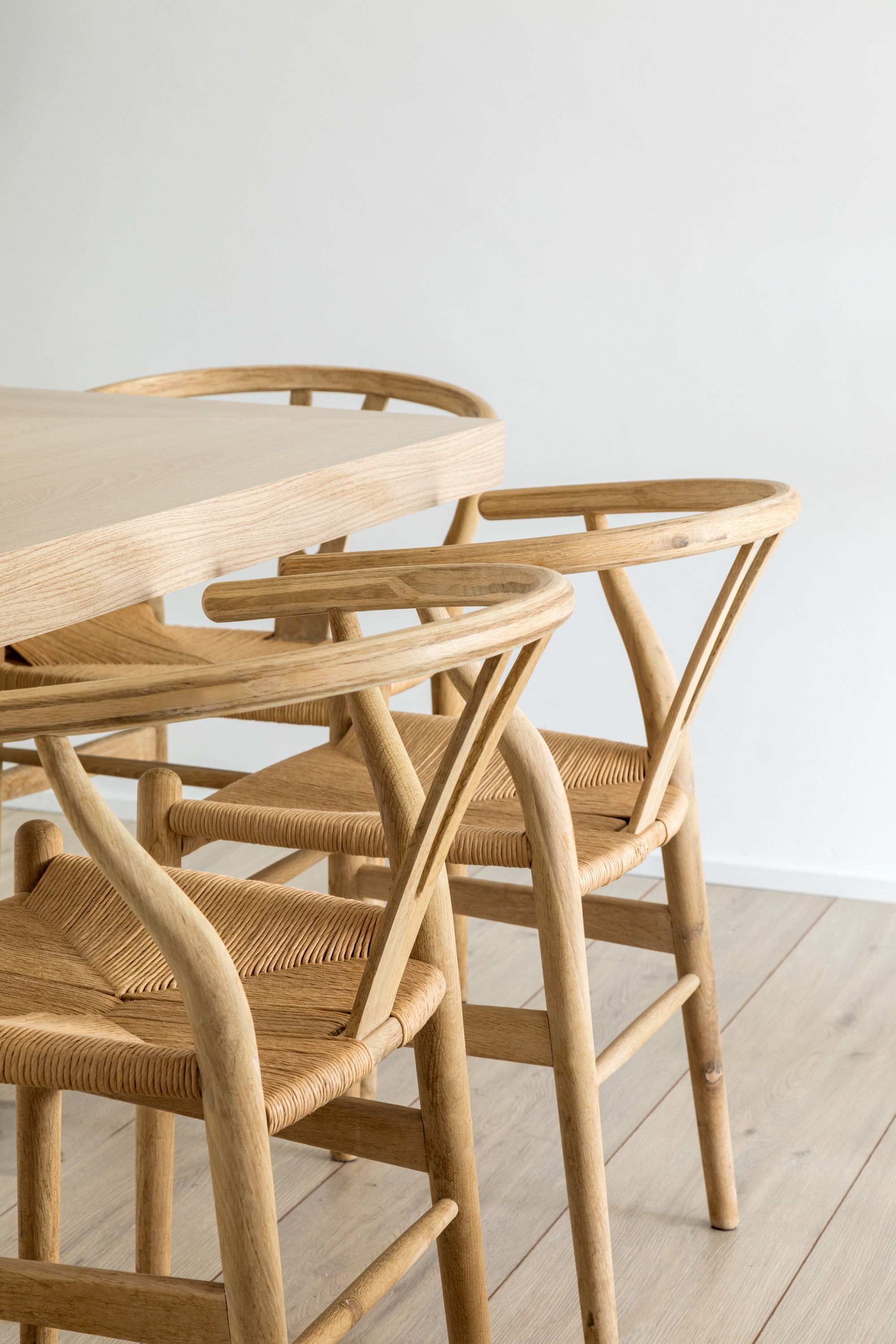 Close-up of woven paper cord seats and curved oak armrests on wishbone chairs beside a light timber table edge