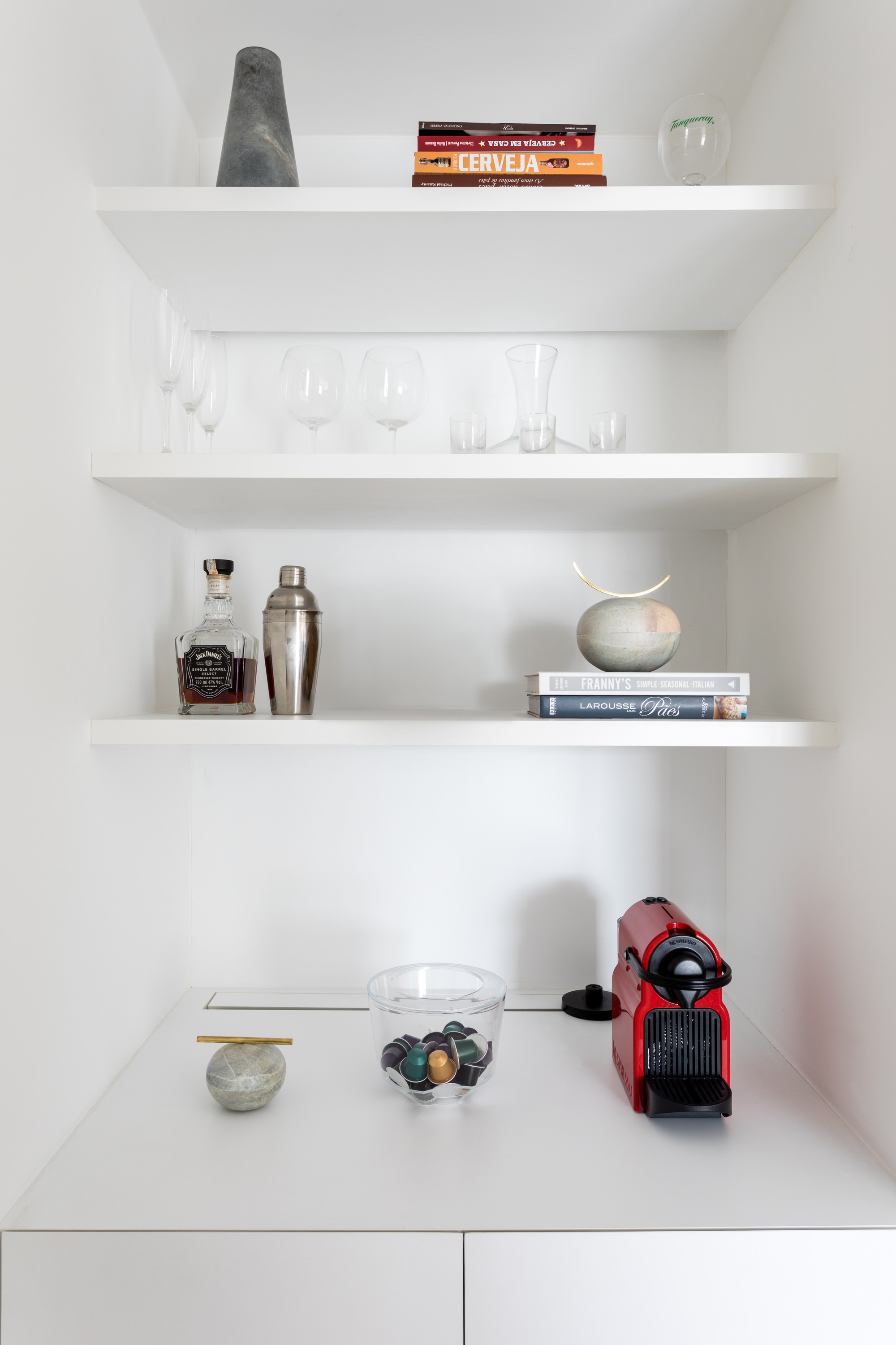 Built-in white shelves holding glassware, a cocktail shaker, books and a red capsule coffee machine on the cabinet below