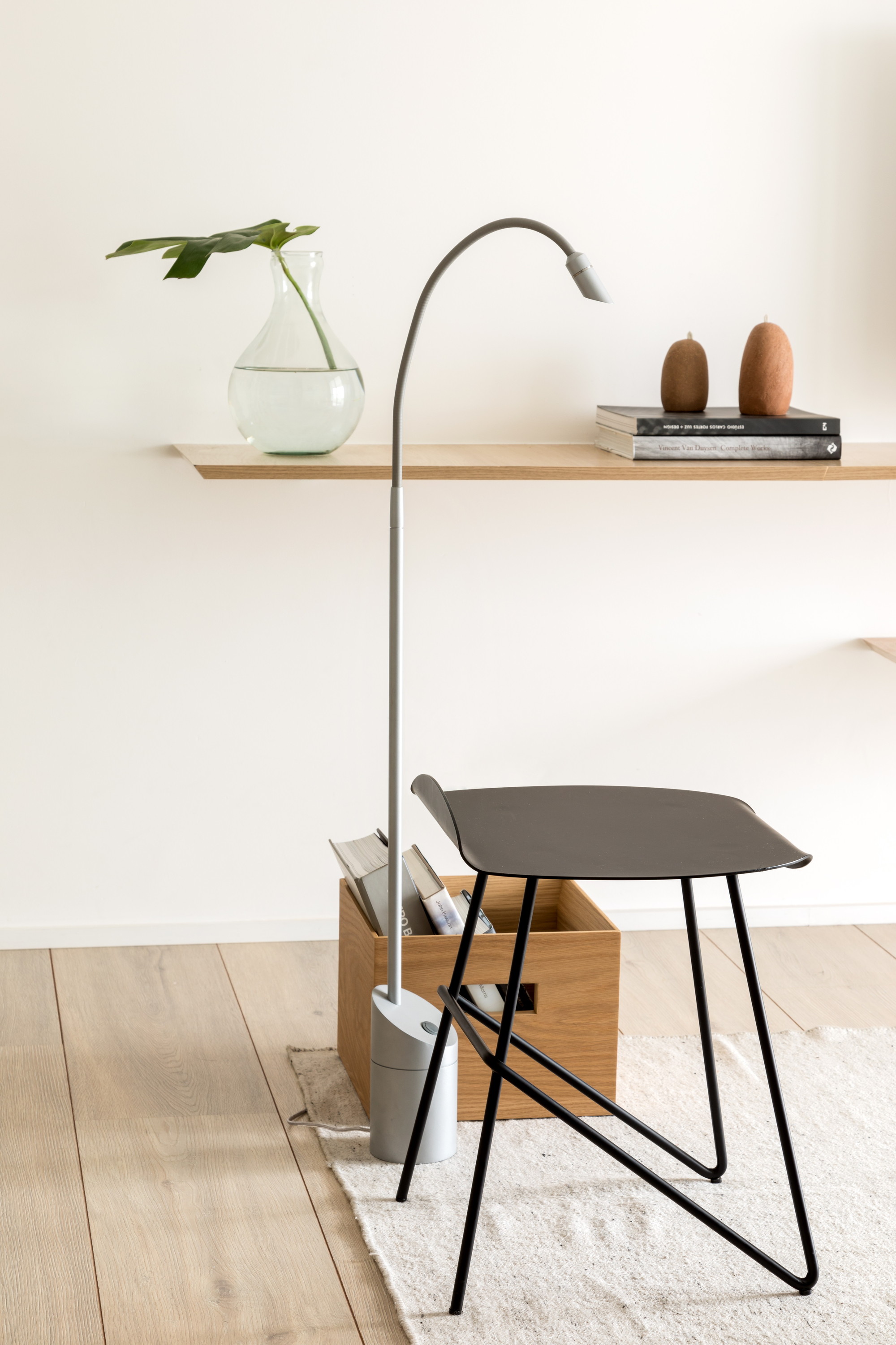 Black metal stool and grey arched floor lamp beside a wooden crate of magazines, with a glass vase on the oak shelf behind