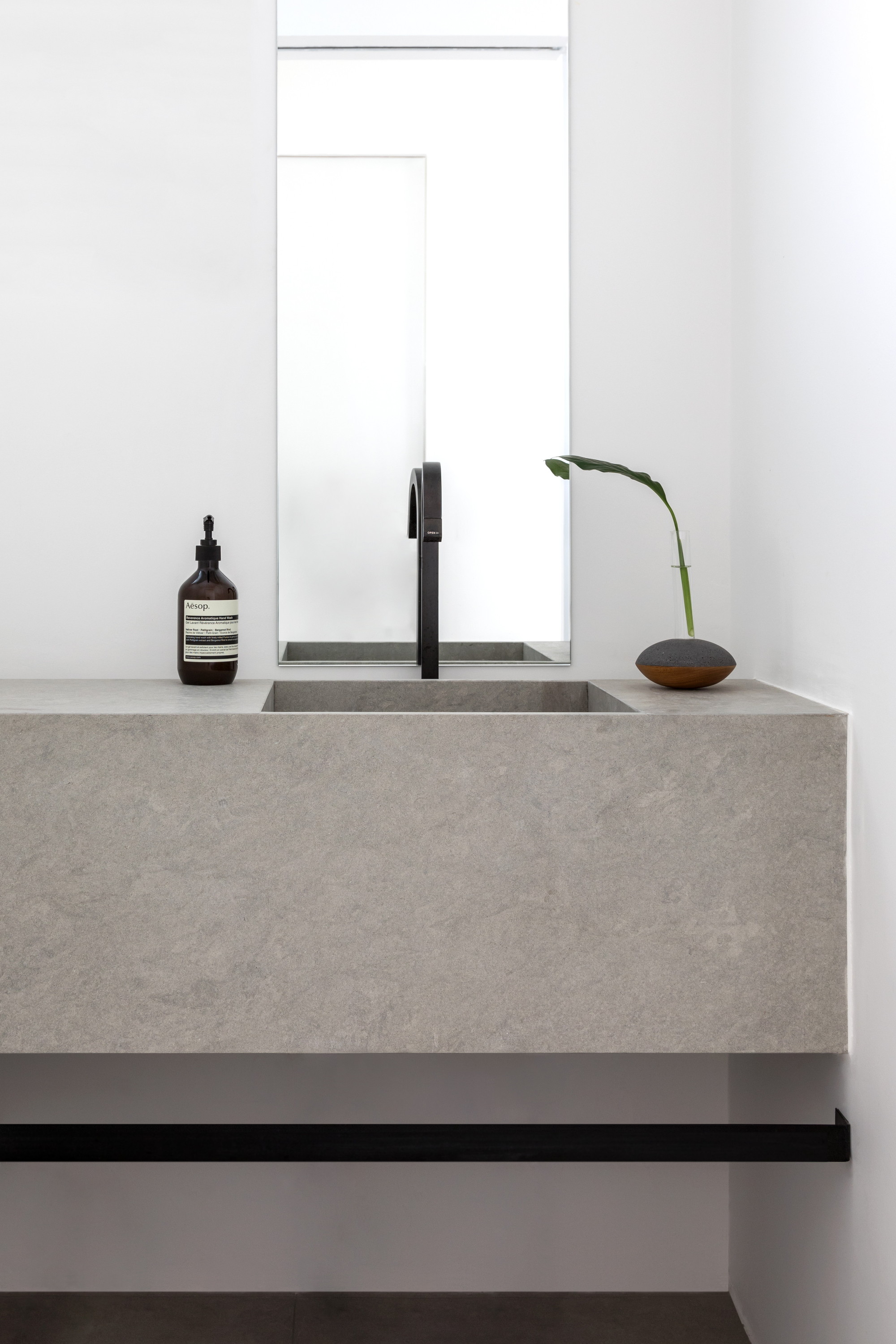 Floating grey stone vanity with integrated basin, black arched tap and a single leaf stem in soft daylight