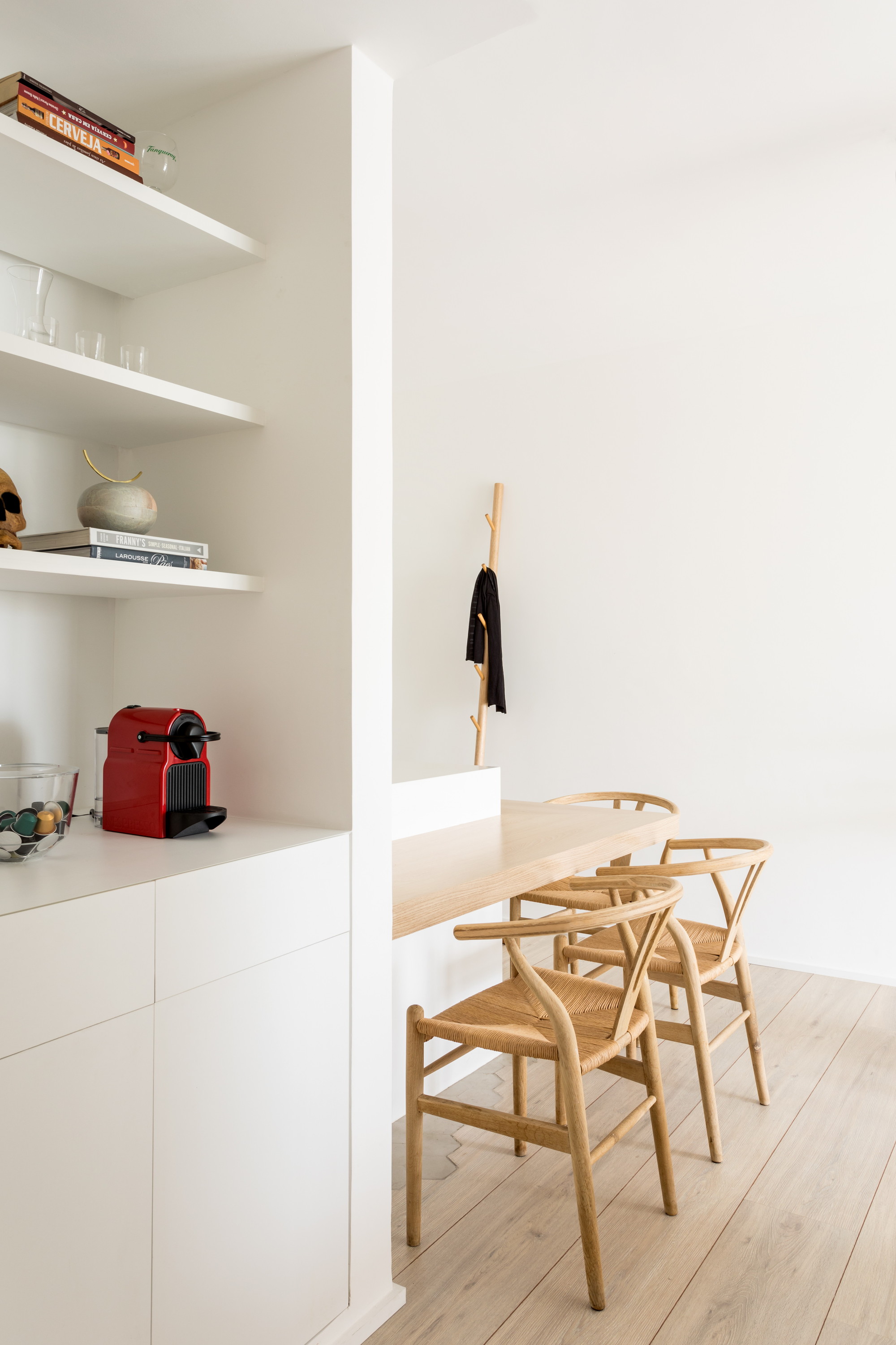 Wishbone chairs at a timber wall-mounted counter beside white shelving and a red coffee machine, with a coat hung on a wooden stand