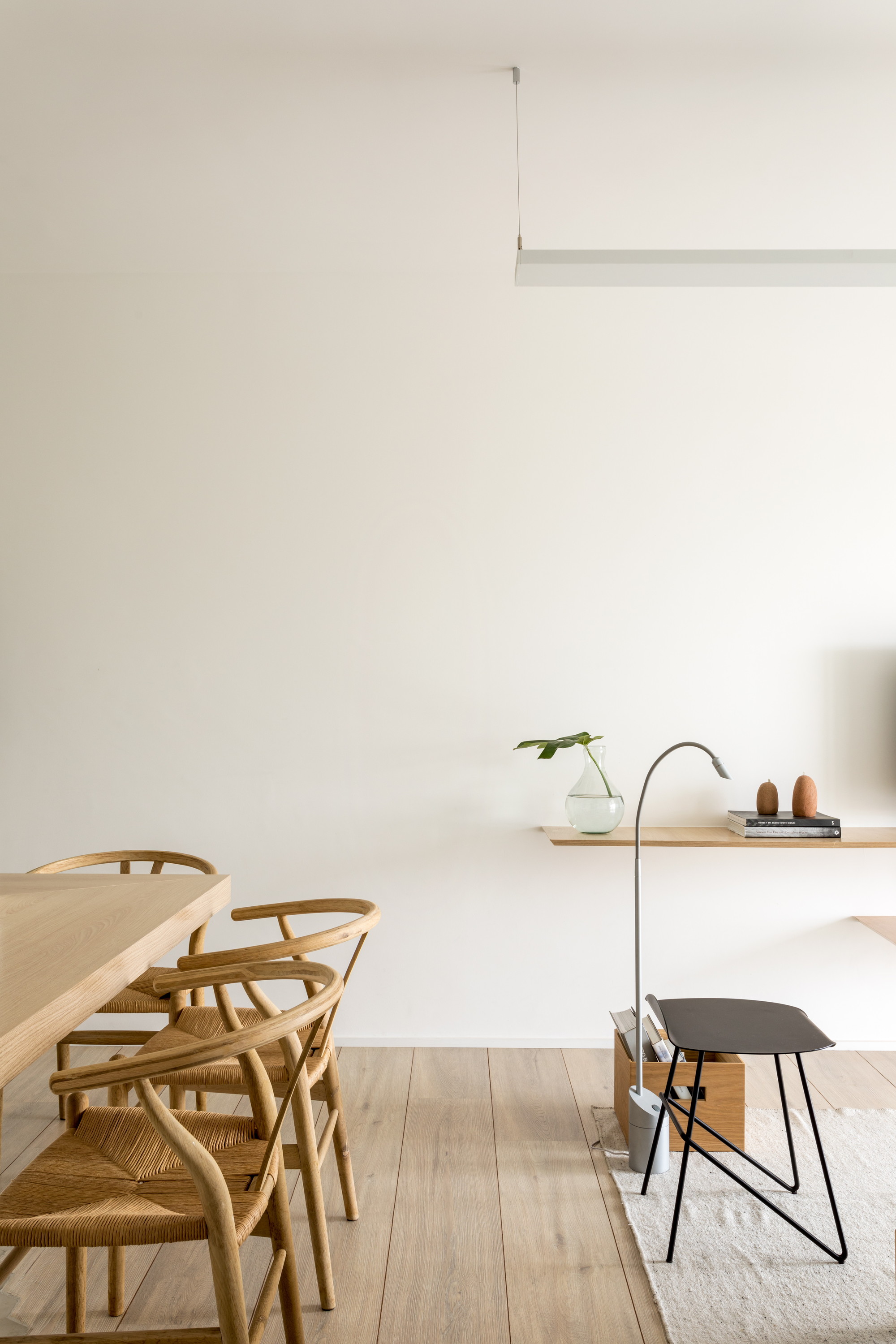 Dining chairs and oak table facing a slim wall shelf, floor lamp and black stool on a pale rug