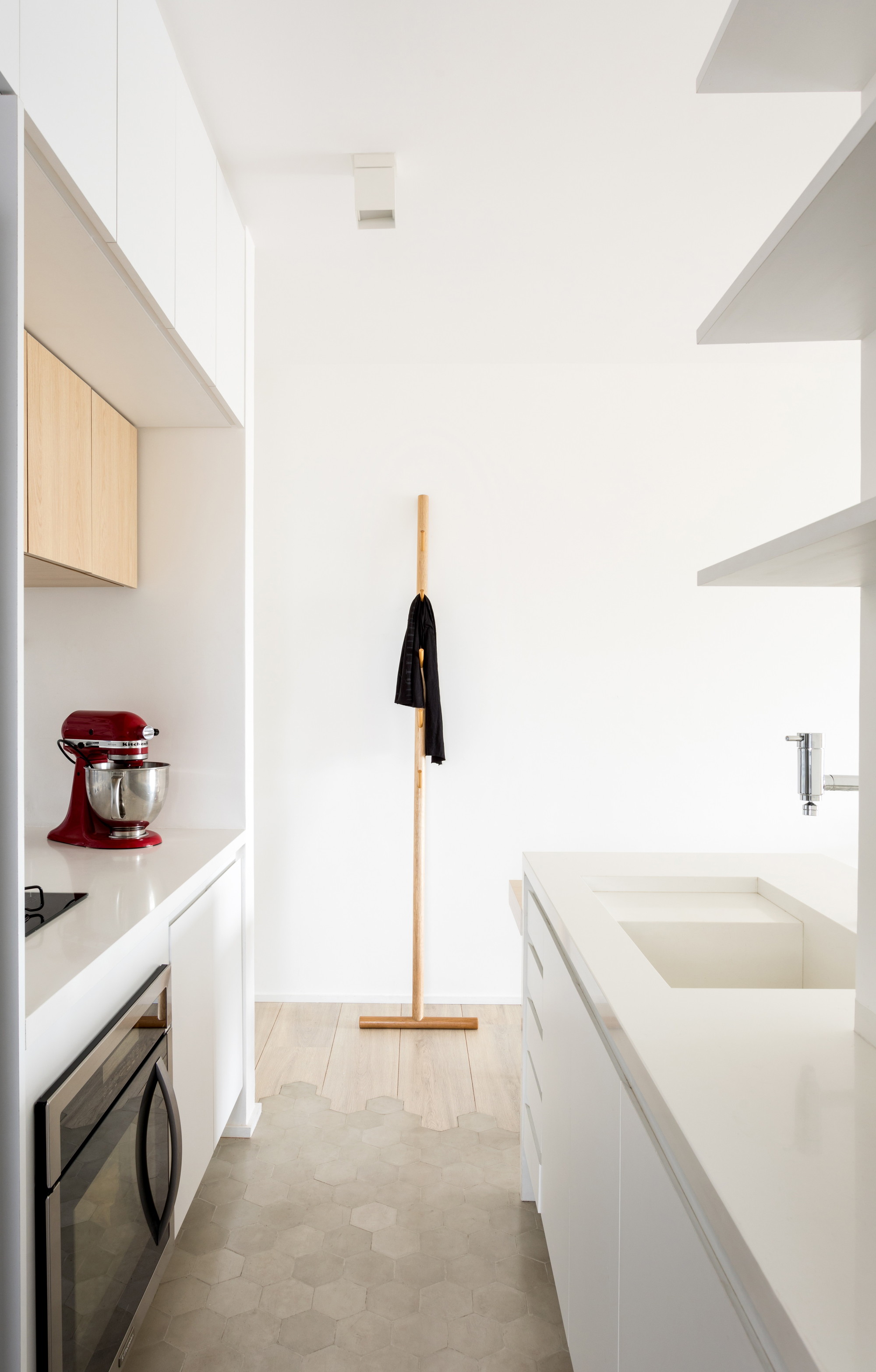 Narrow white kitchen with a red stand mixer, hexagonal floor tiles and a wooden coat stand at the far end