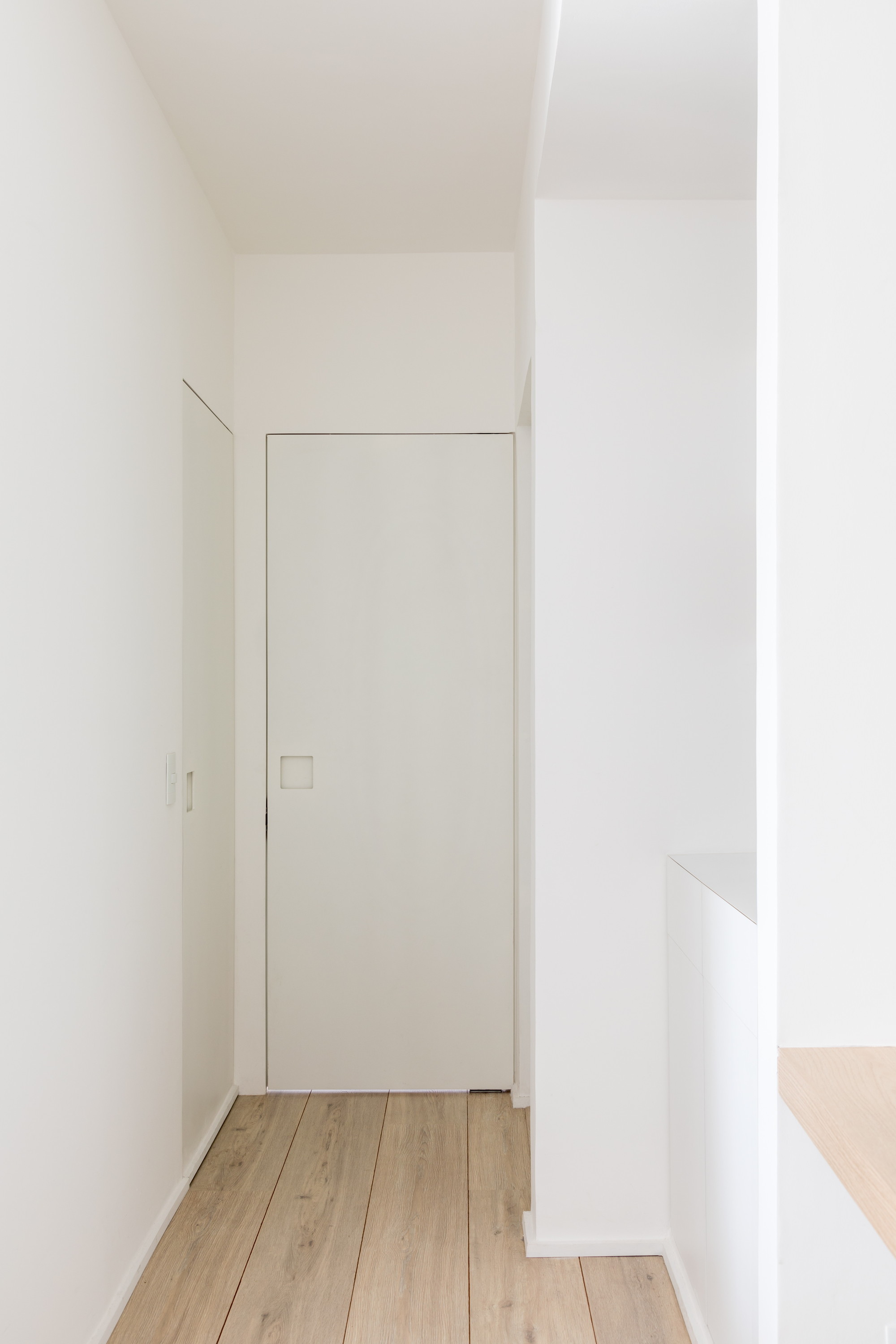 Flush white door with a square cut-out handle at the end of a short hallway of pale plank flooring