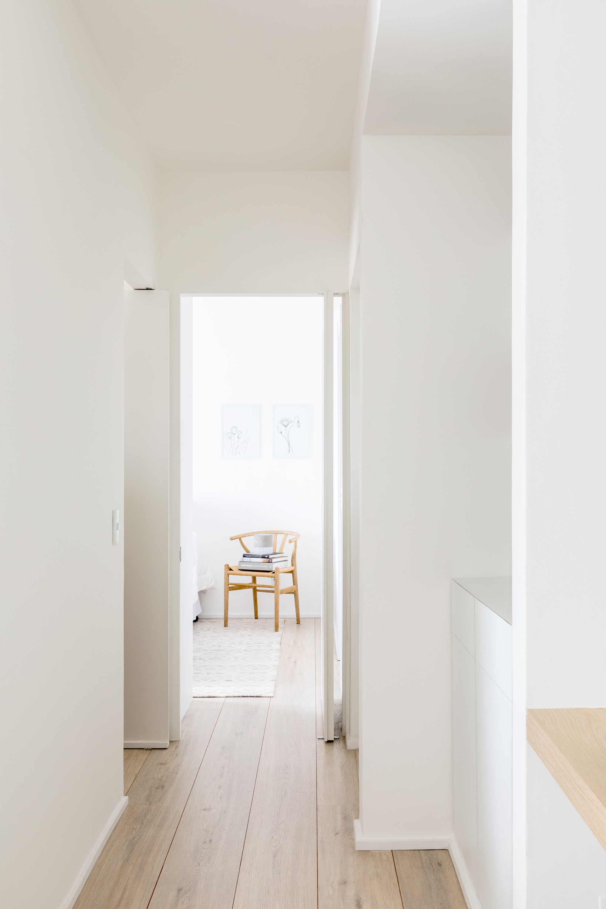 Narrow white hallway with pale oak floorboards leading to a wooden Wishbone-style chair and framed botanical prints in soft daylight