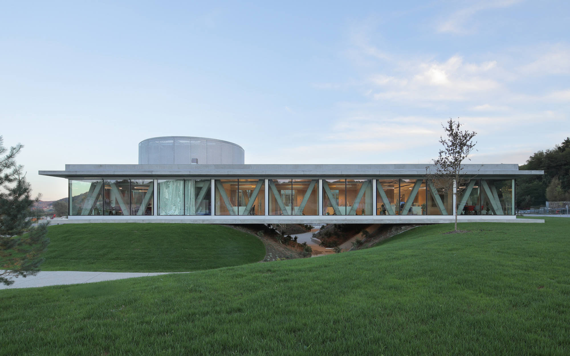 Long glazed pavilion with diagonal concrete bracing and a cylindrical volume above, set on a grassy slope at dusk