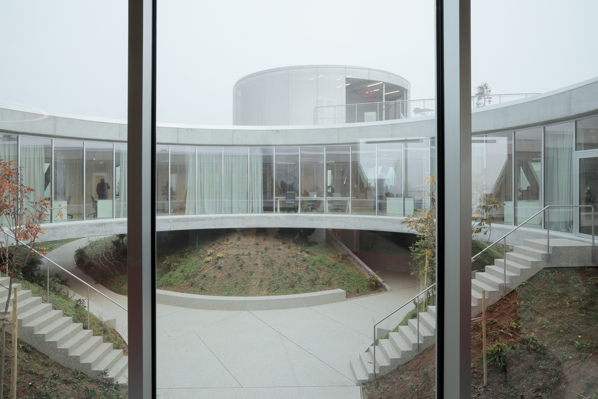 Curved ring-shaped building around a sunken courtyard with planted mound and concrete stairs, seen through a window in fog
