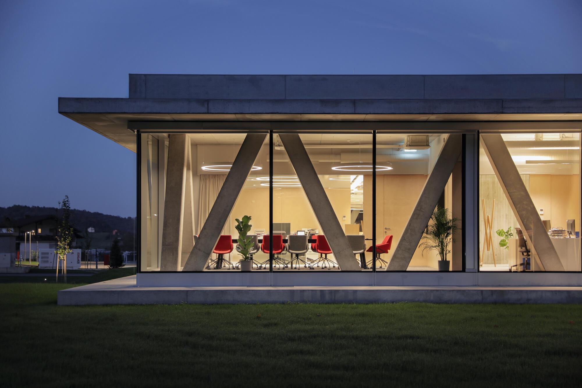 Glazed end of the building with diagonal concrete struts and red chairs around a meeting table, lit at dusk