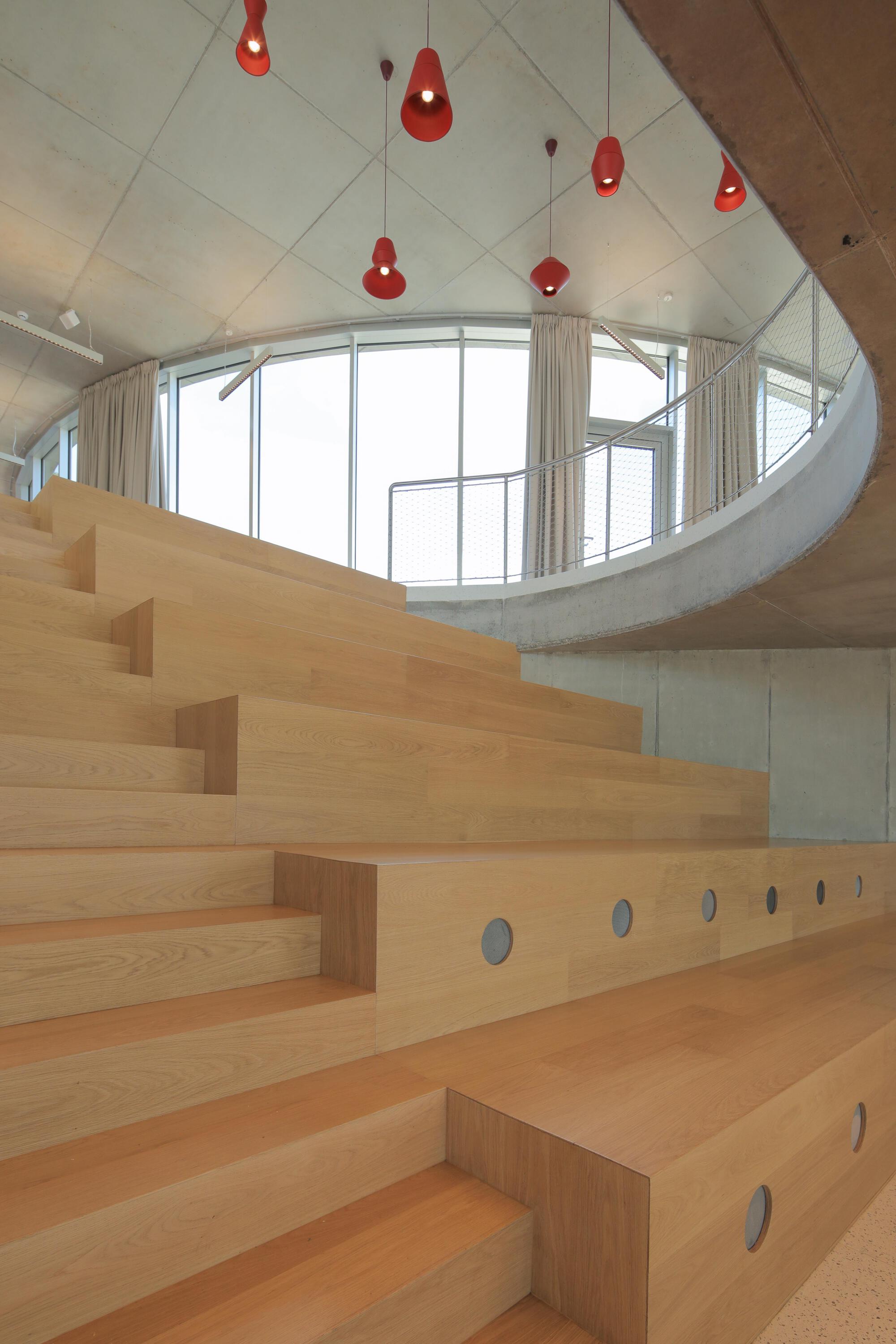 Timber stepped seating with round cutouts beneath a curved window wall and red pendant lamps, soft daylight through curtains