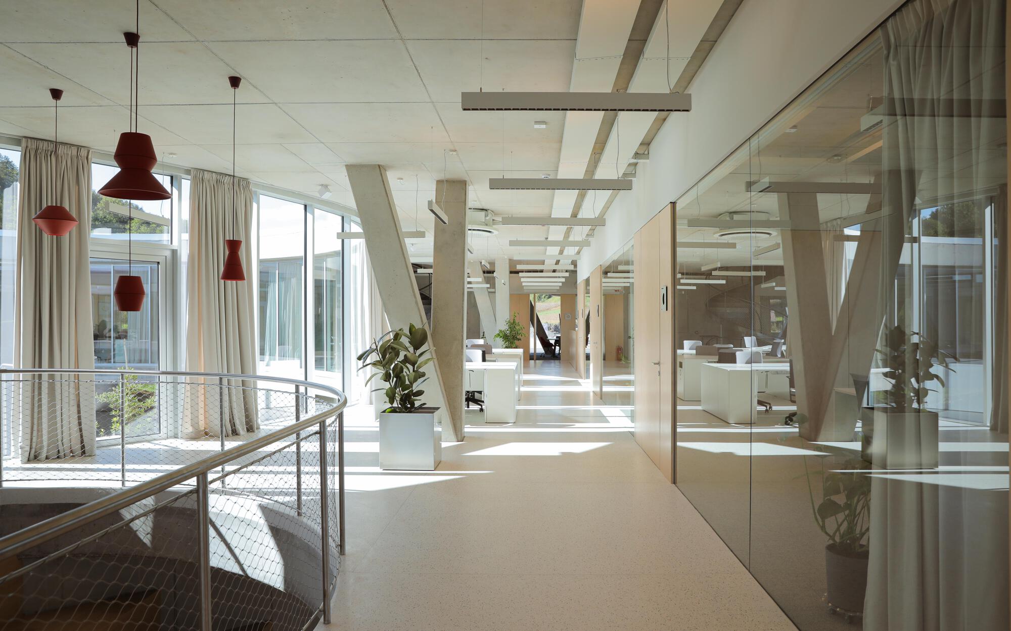 Open office corridor with slanted concrete columns, mesh balustrade and red pendant lamps in bright sunlight