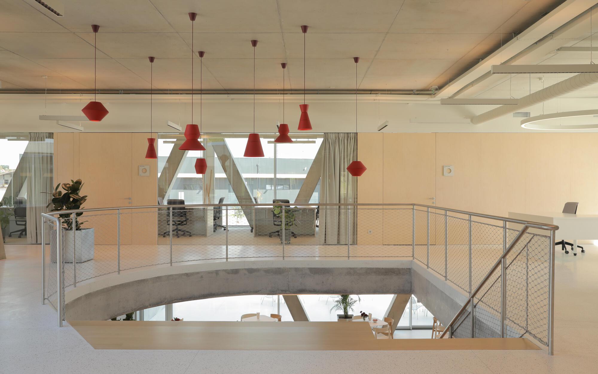 Open stair void with a mesh balustrade and red pendant lamps hanging below a concrete ceiling, daylight filtering in from the glazed facade
