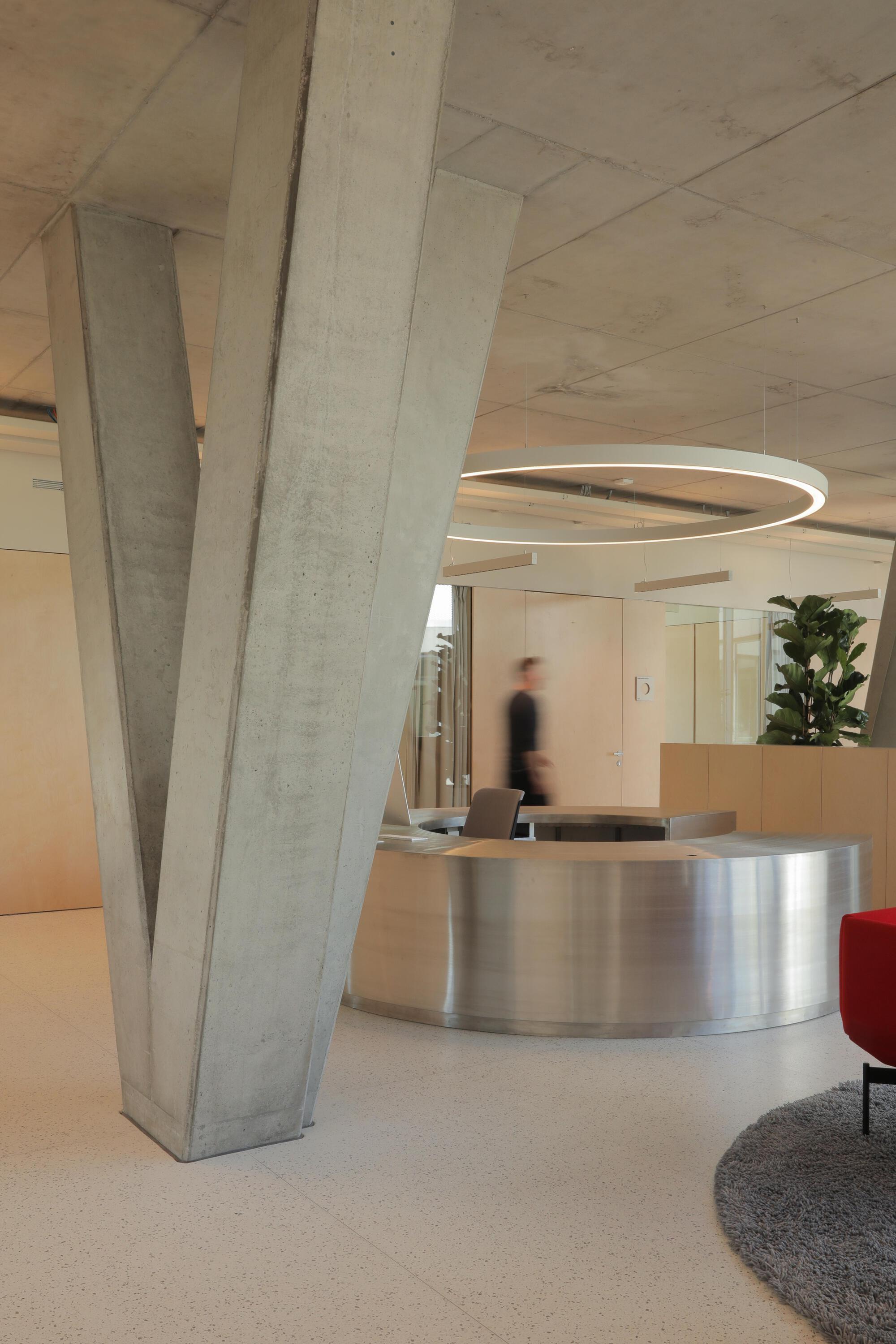 Forked concrete column beside a curved stainless steel reception desk, with a person blurred in motion behind and a ring light overhead