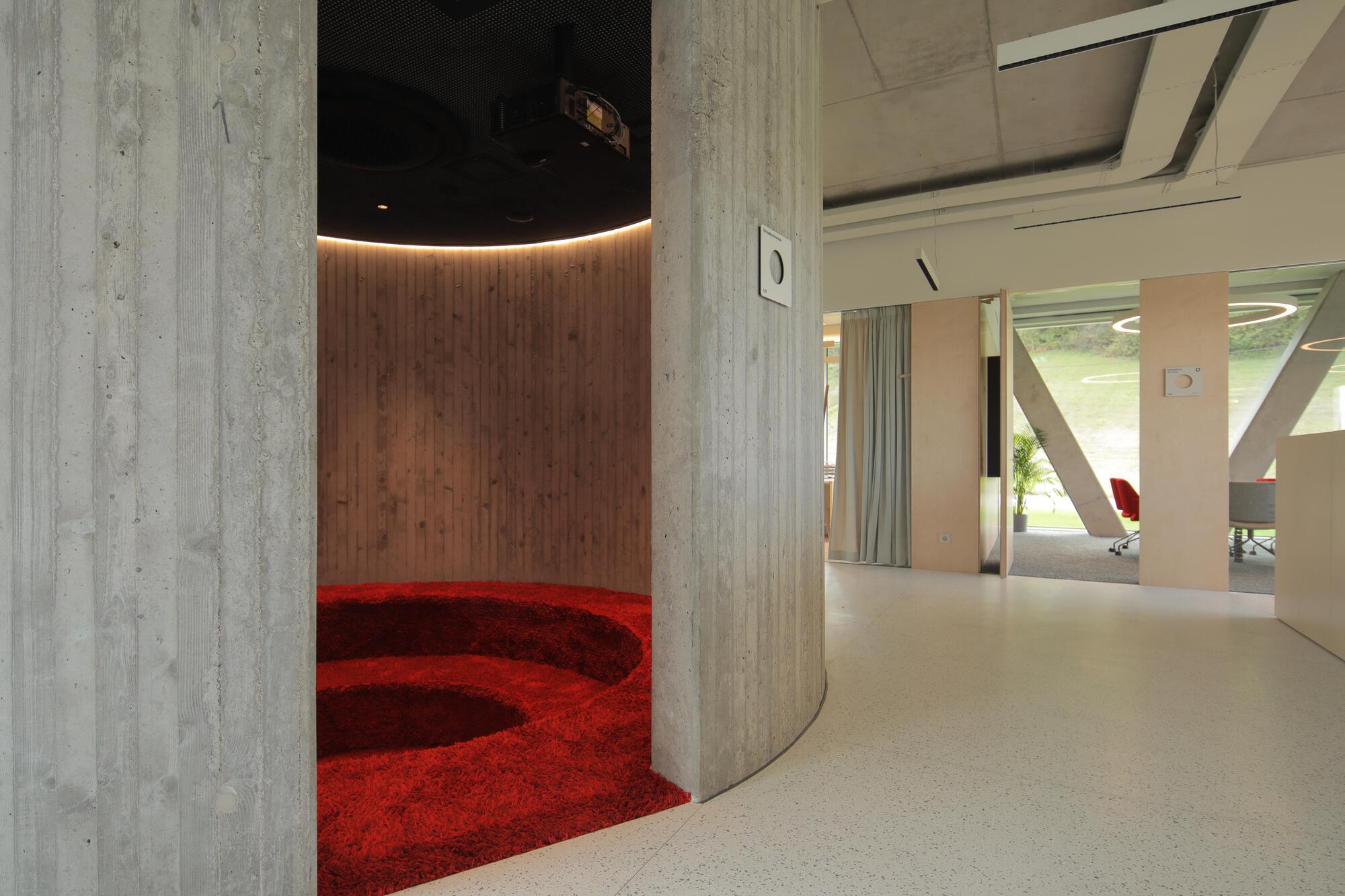 Sunken seating pit with red shag carpet inside a curved board-formed concrete enclosure, lined with timber and lit by a warm cove light