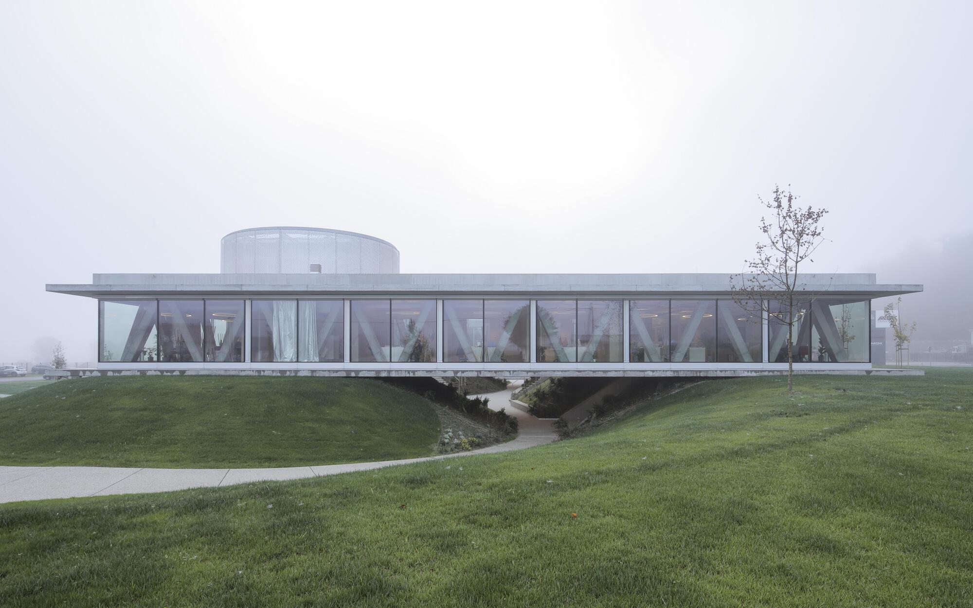 Long glazed pavilion with diagonal concrete bracing, raised over a grass mound and a curving path, under dense fog