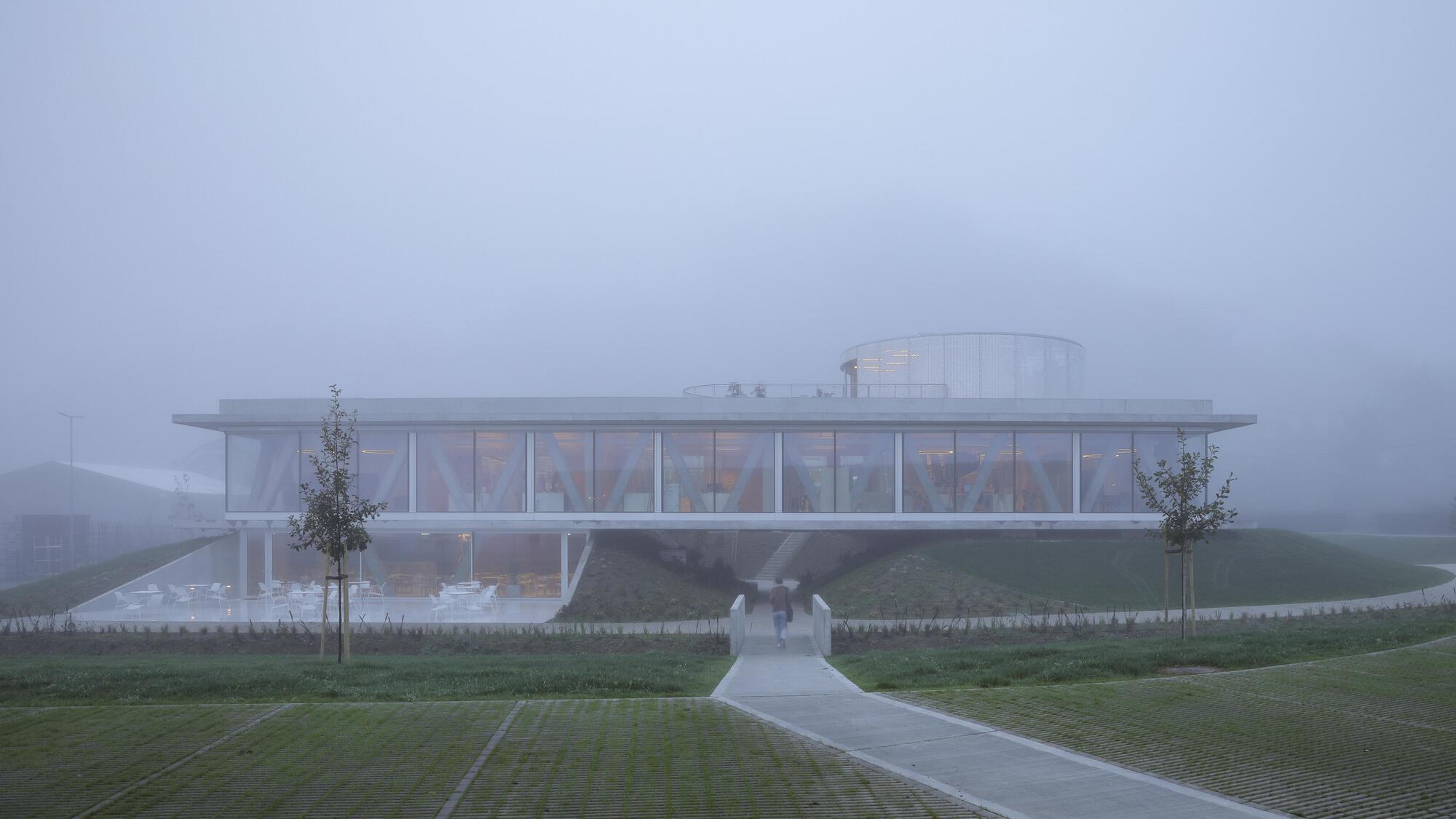 Glazed pavilion with a cylindrical rooftop volume seen across paving and lawn, a lone figure walking the path in thick mist