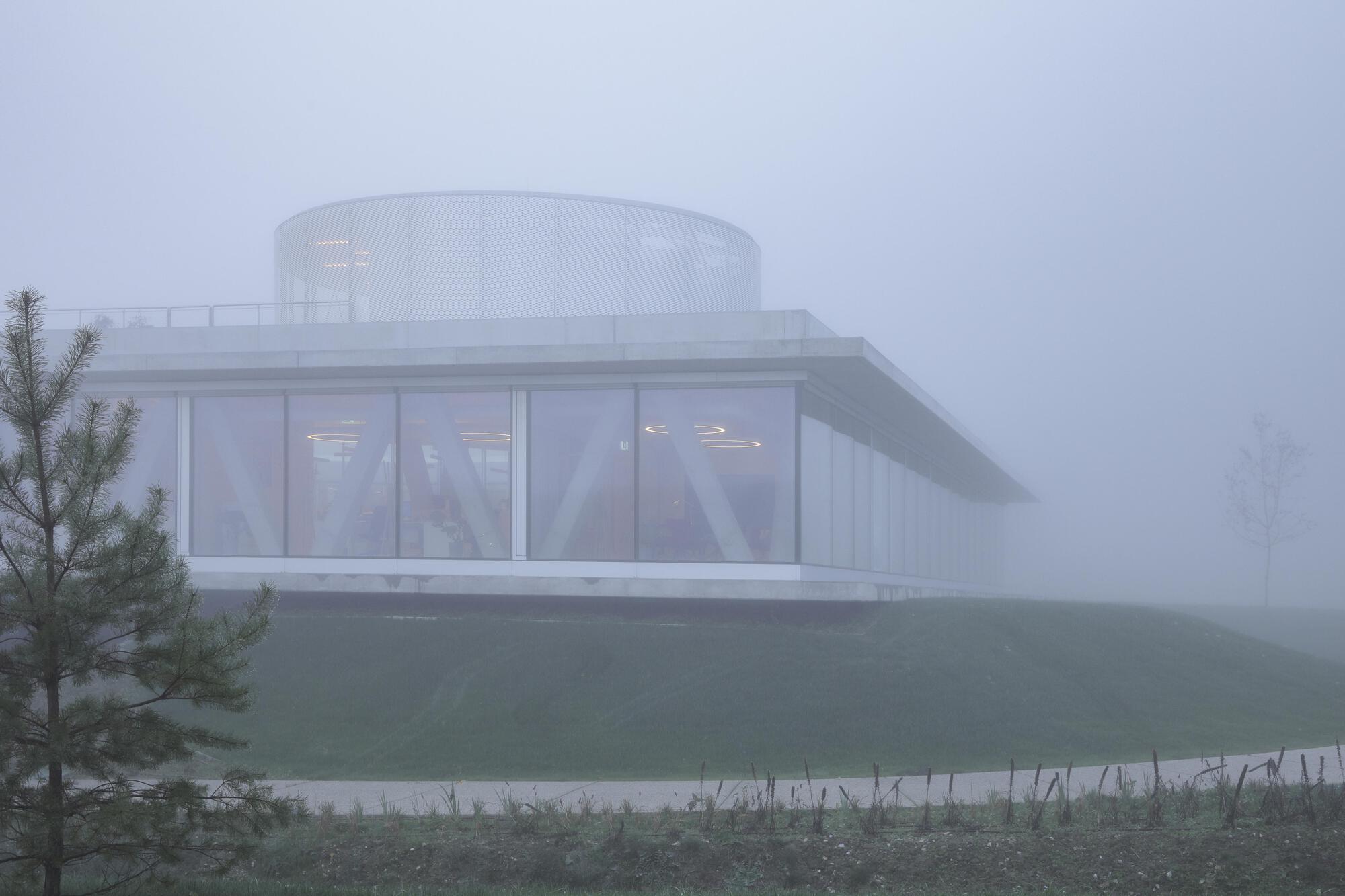 Glazed pavilion with a cylindrical rooftop volume and cantilevered concrete roof, seen across a grassy slope in dense fog