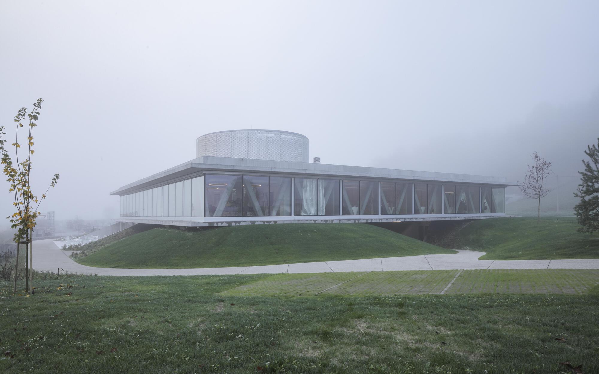 Low glass pavilion with diagonal interior struts and a round rooftop drum, resting on a green mound under fog
