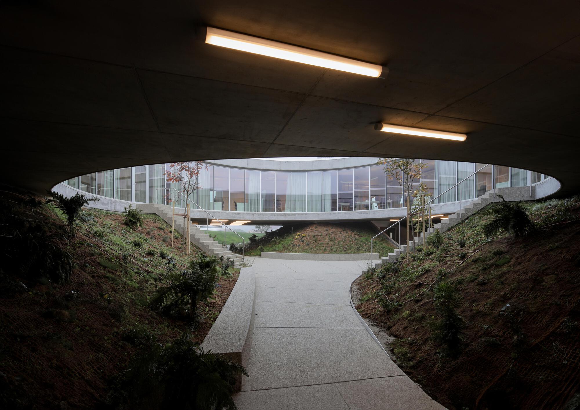 View from beneath a concrete underpass with strip lights, out to a sunken courtyard path, stairs and a glazed ring building
