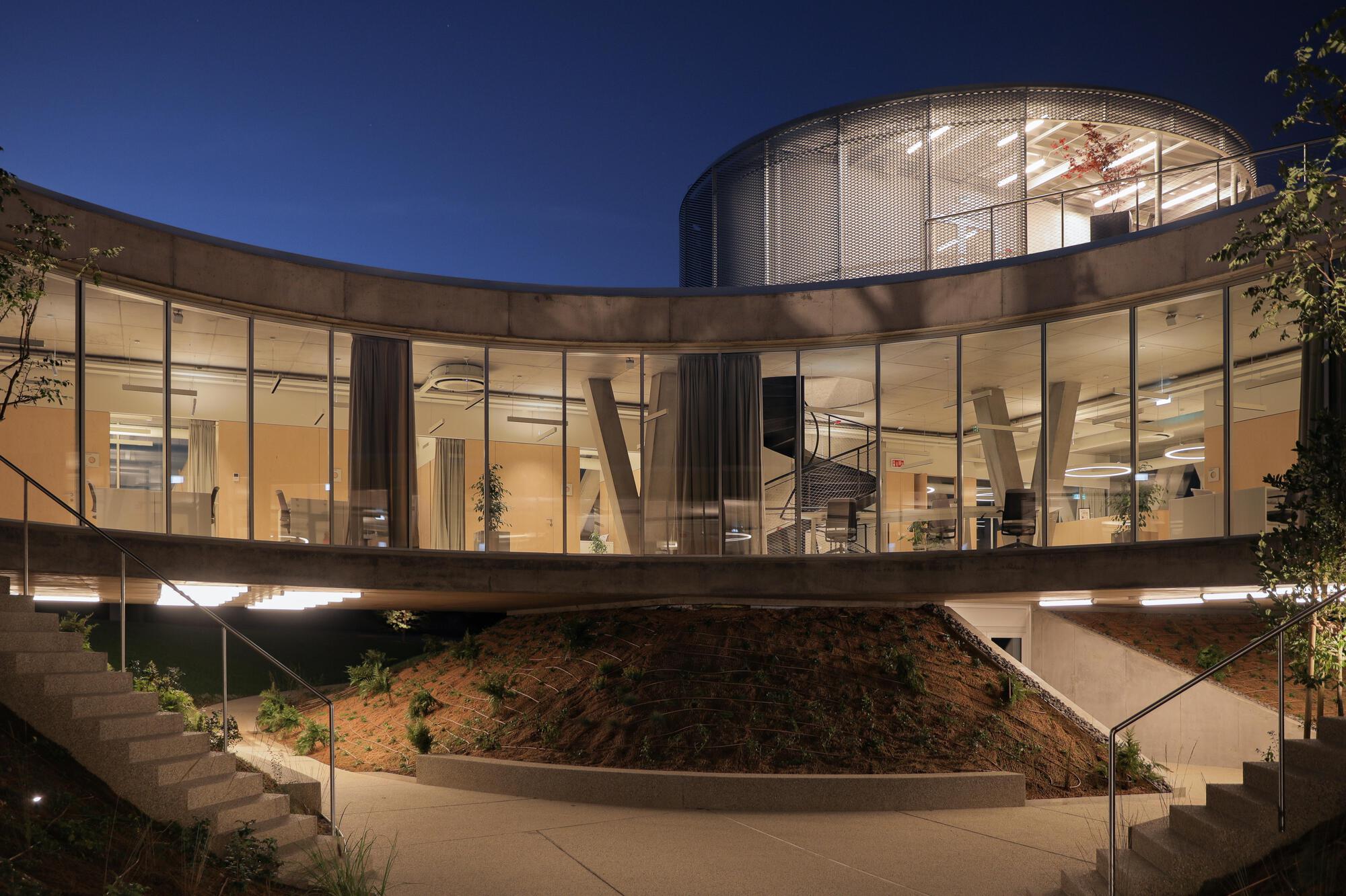 Dusk view of the curved glass office level with angled columns and a mesh-clad drum above, lit from within