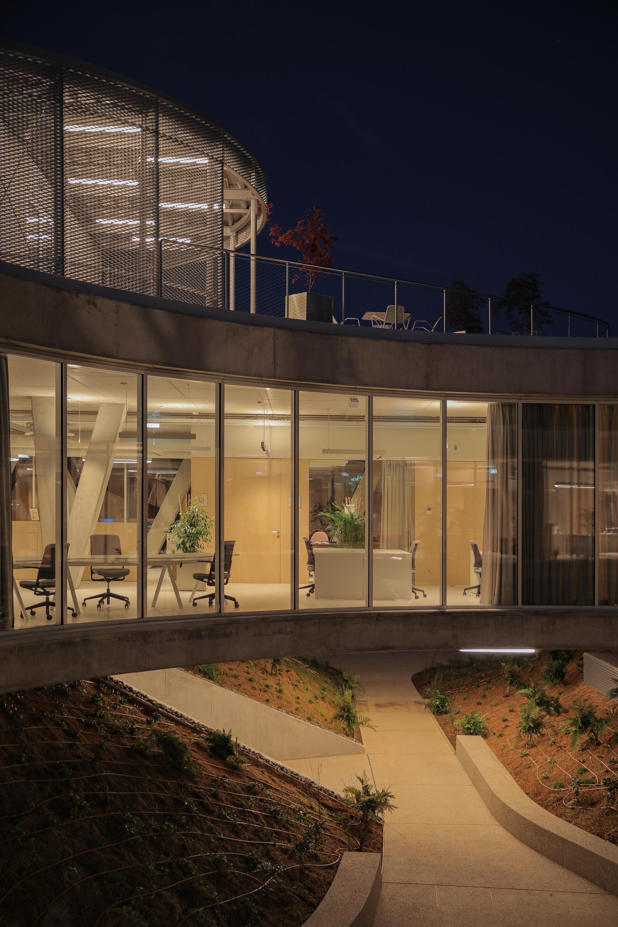 Curved glass wall of an office floor above a sunken planted courtyard, lit from within at night with a rooftop terrace overhead