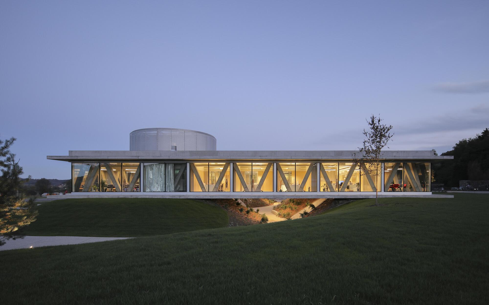 Long elevation of a glazed pavilion with diagonal timber bracing and a cylindrical drum on the roof, seen over lawn at dusk