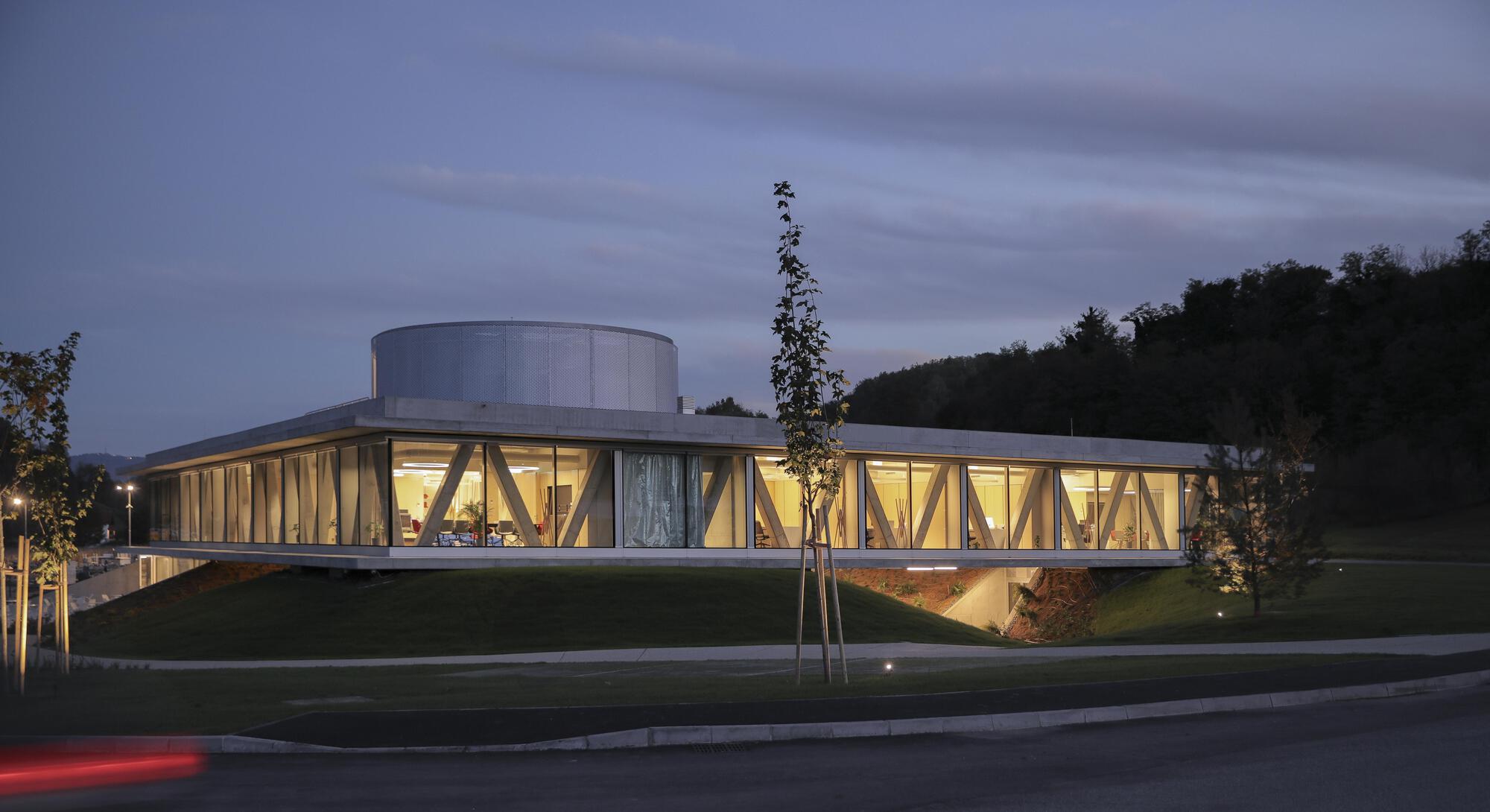 Corner view of the cantilevered glass-walled volume and roof drum above grassy mounds, with a young tree and a car light streak at dusk
