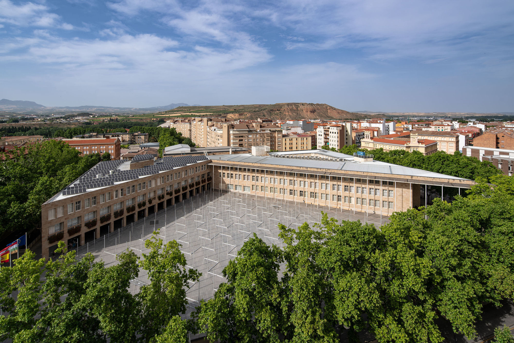 Aerial view of a stone-clad courtyard building with solar panels on its roof, ringed by green plane trees under scattered clouds