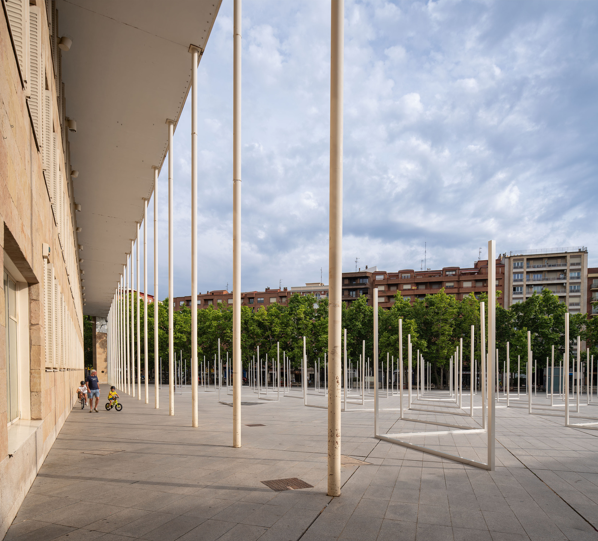 Slender white poles rising from the paving beside a long stone colonnade, with two children cycling past under a cloudy sky