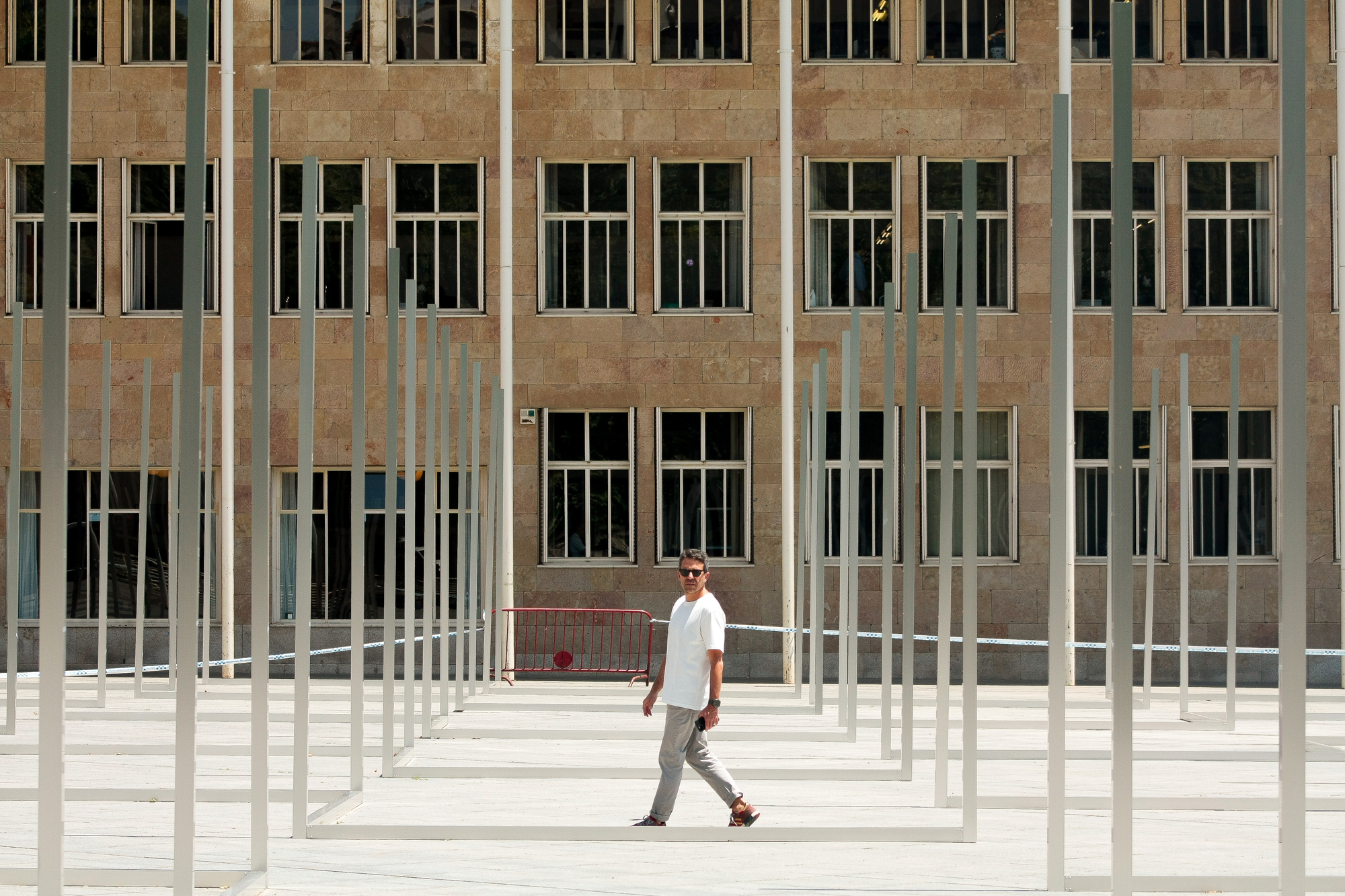 Pedestrian walking between thin steel uprights and floor-level frames in front of a stone facade with grid windows in bright sun