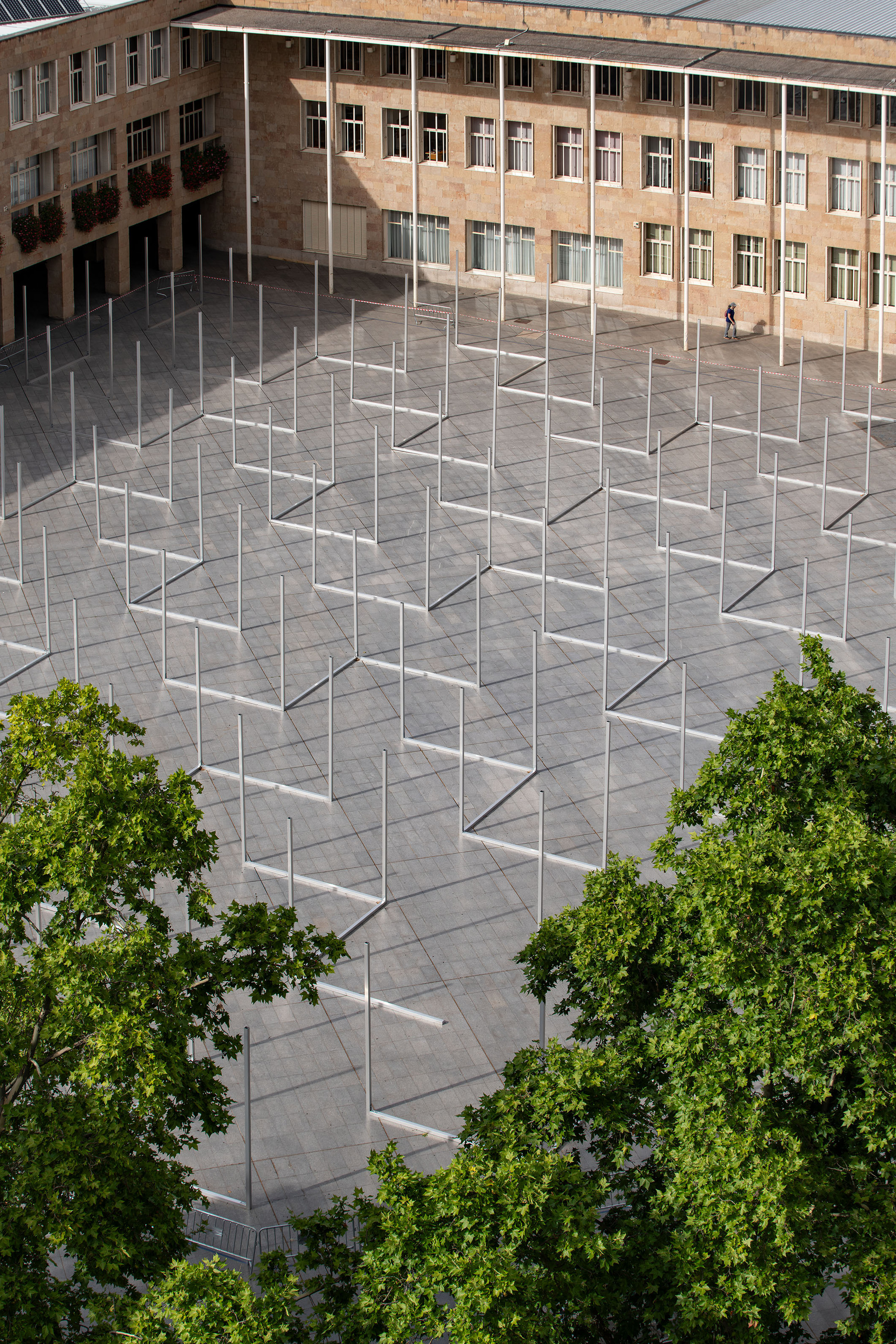 High view of the paved square with rows of steel poles and L-shaped frames, framed by tree canopies and a lone walker