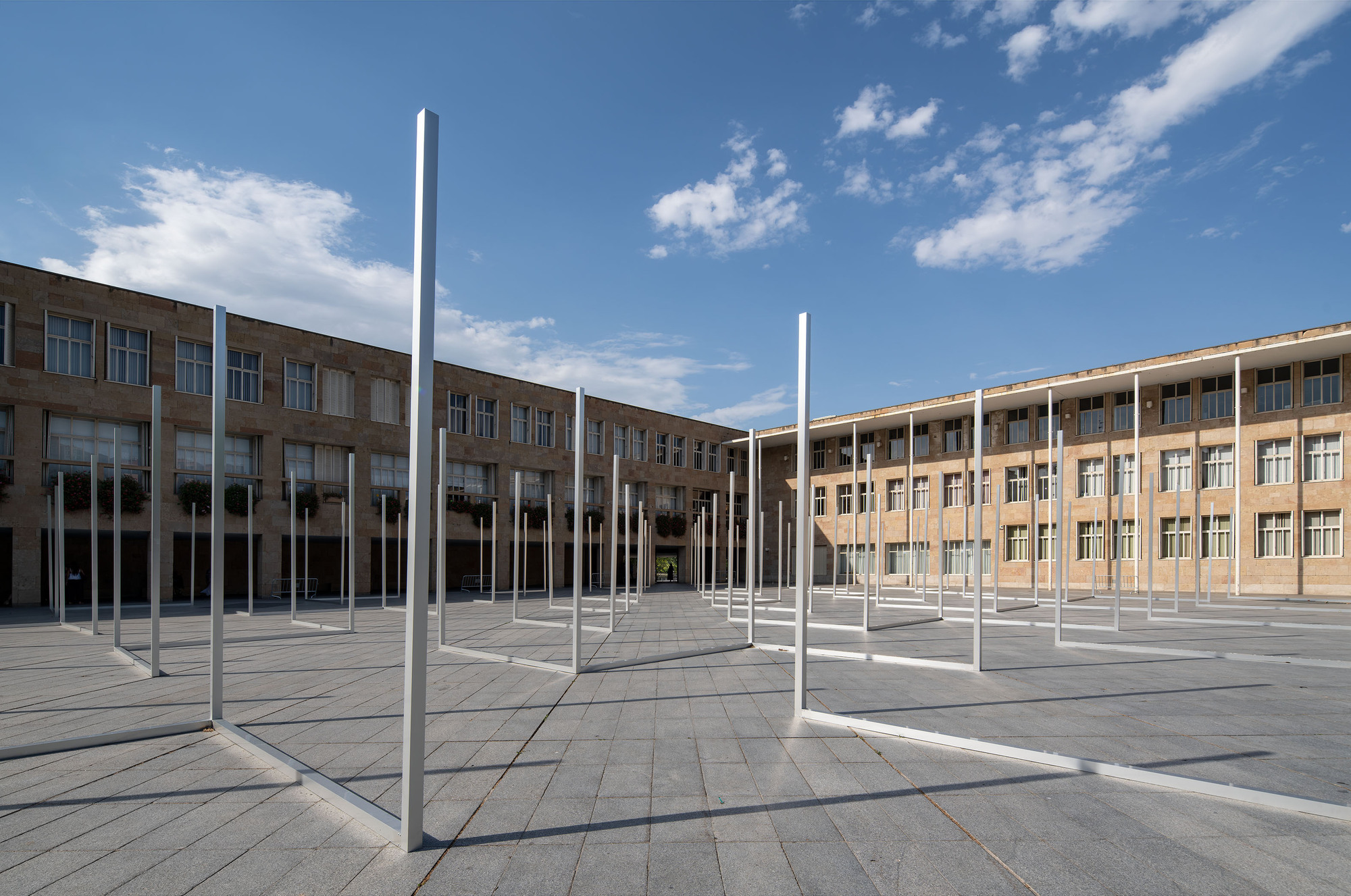 Tall steel poles and flat frame outlines on the paved square, with stone wings behind and a blue sky with clouds