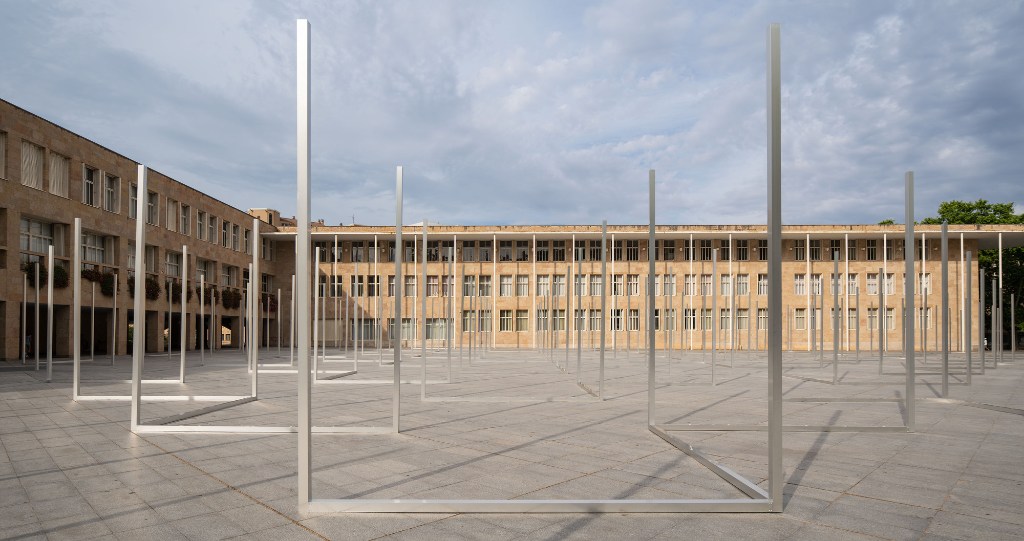 Open rectangular steel frames standing on the paving in front of a long stone building under a soft overcast sky