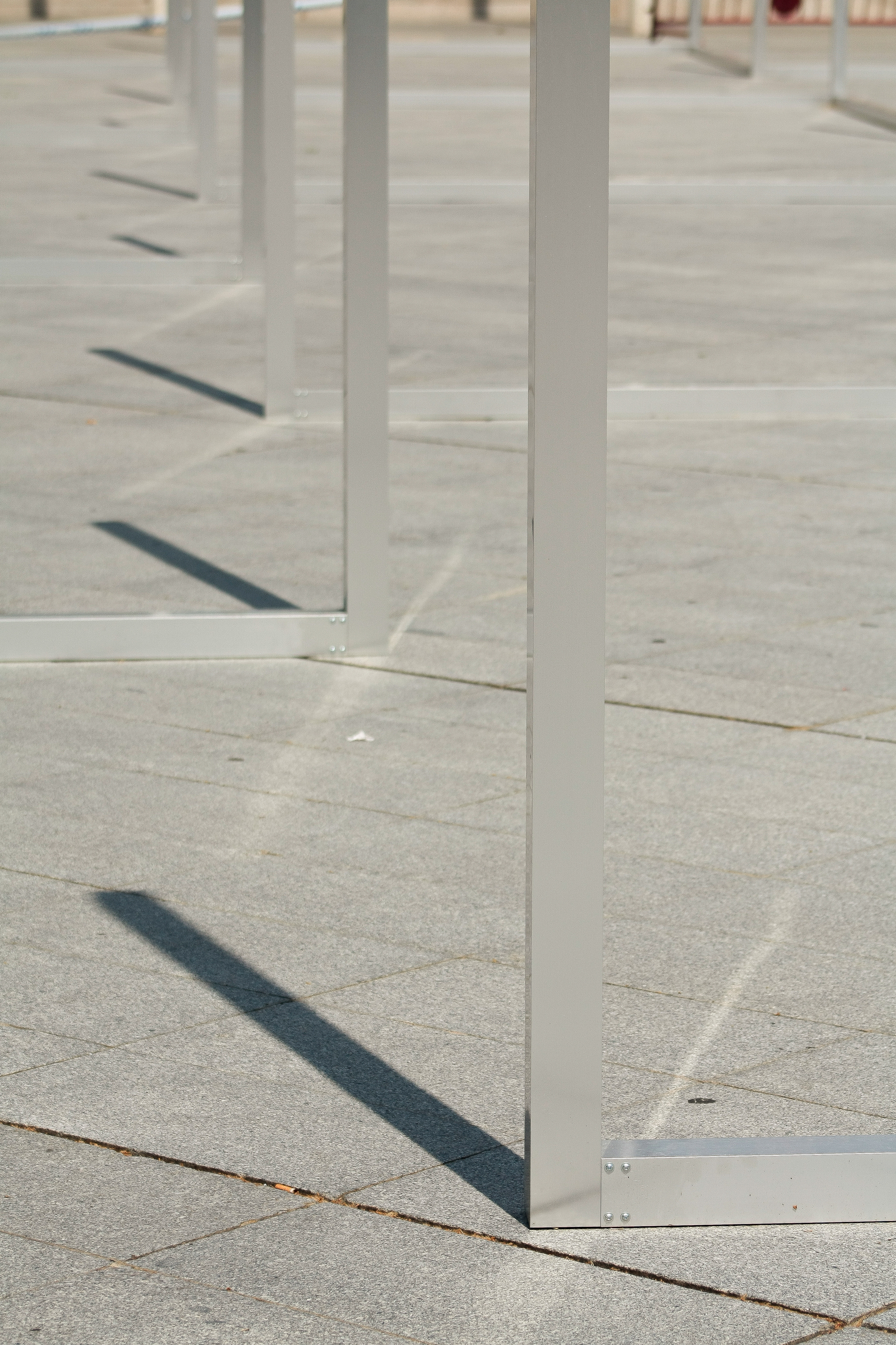 Close-up of slender aluminium frames standing on granite paving, their long diagonal shadows falling across the stone in hard sunlight