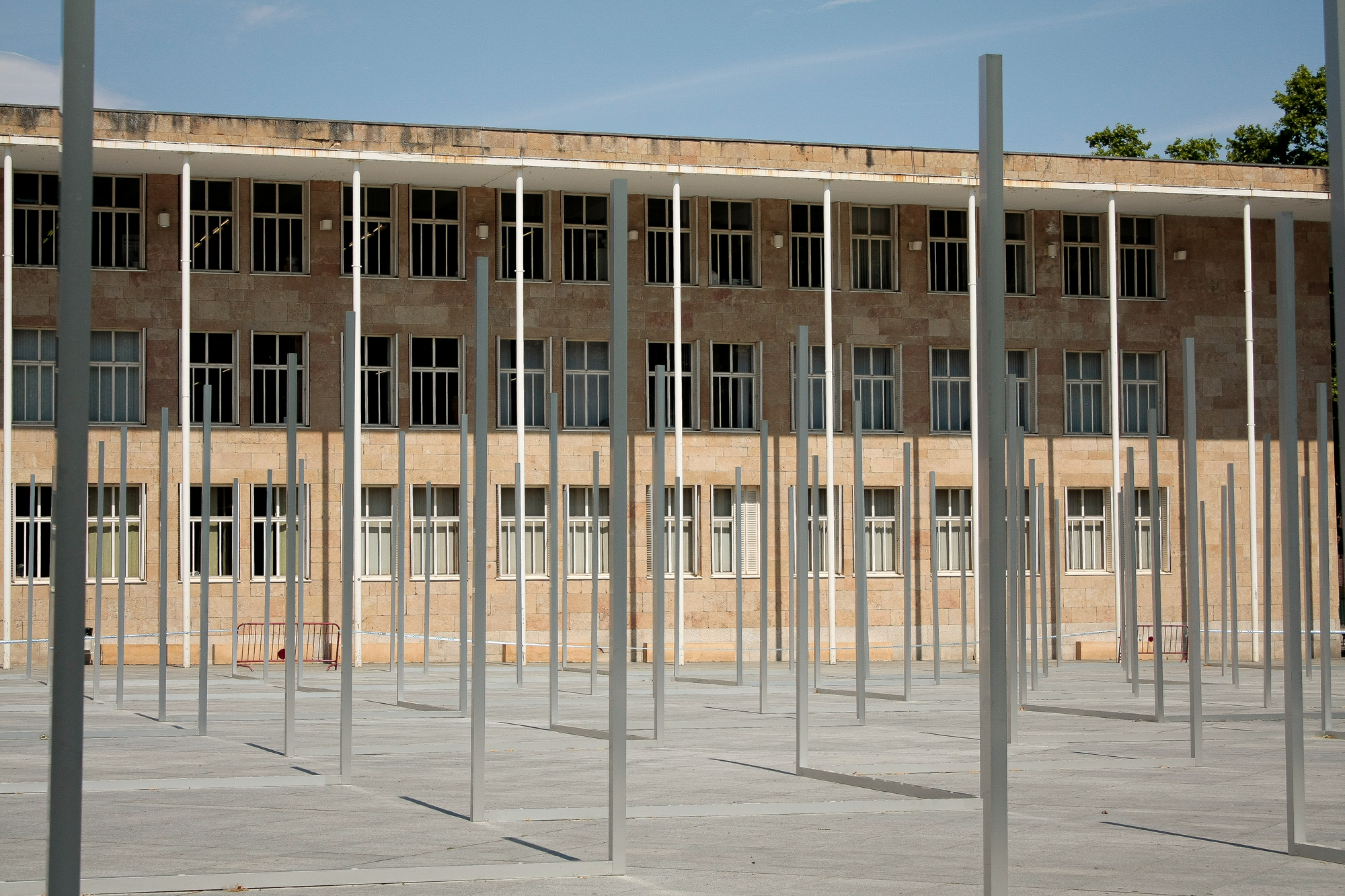 Forest of thin metal posts in a paved plaza in front of a stone-clad office block with rows of windows