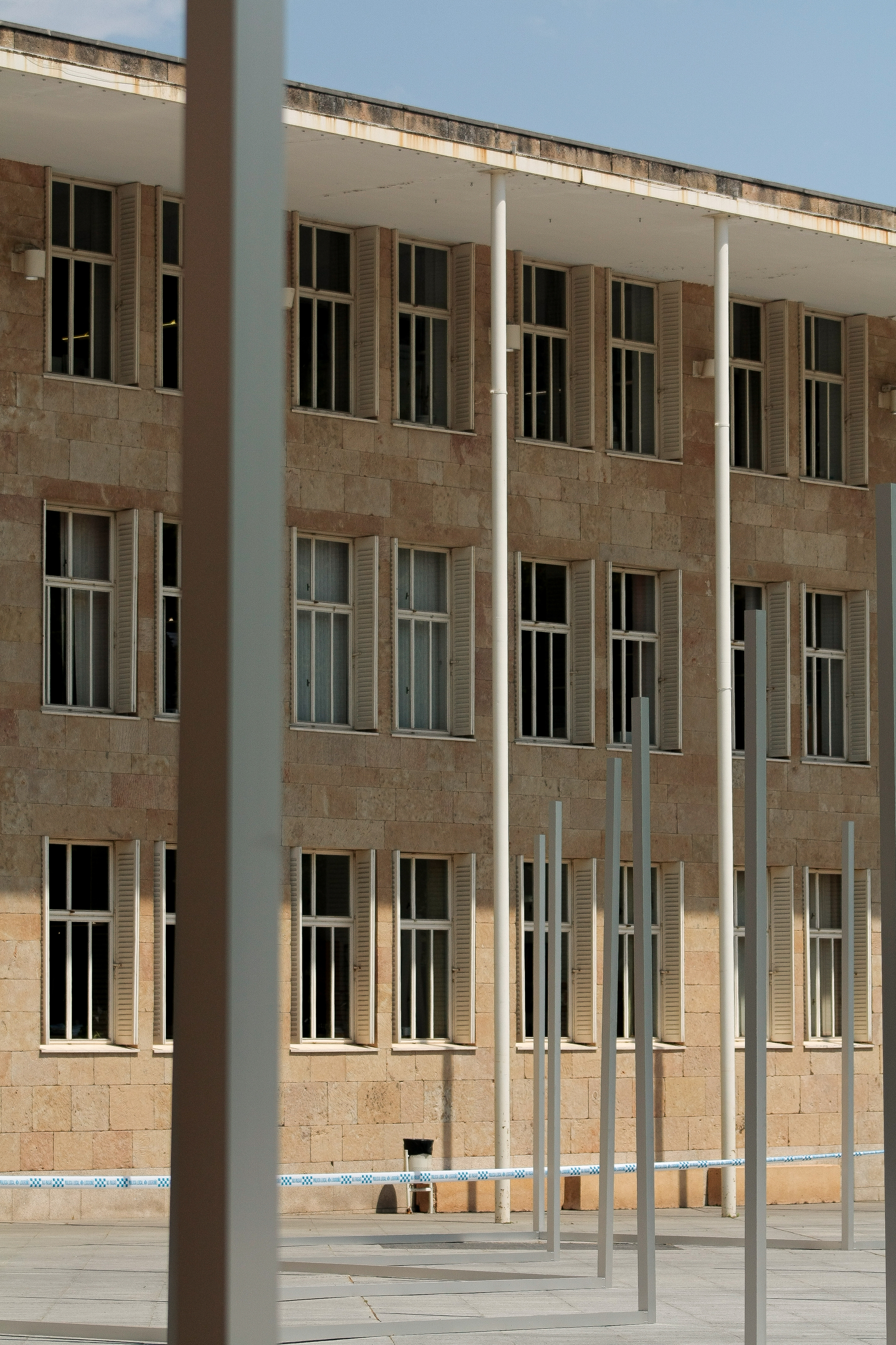 Stone facade with shuttered windows and white columns seen through blurred metal uprights in the foreground, under a pale blue sky