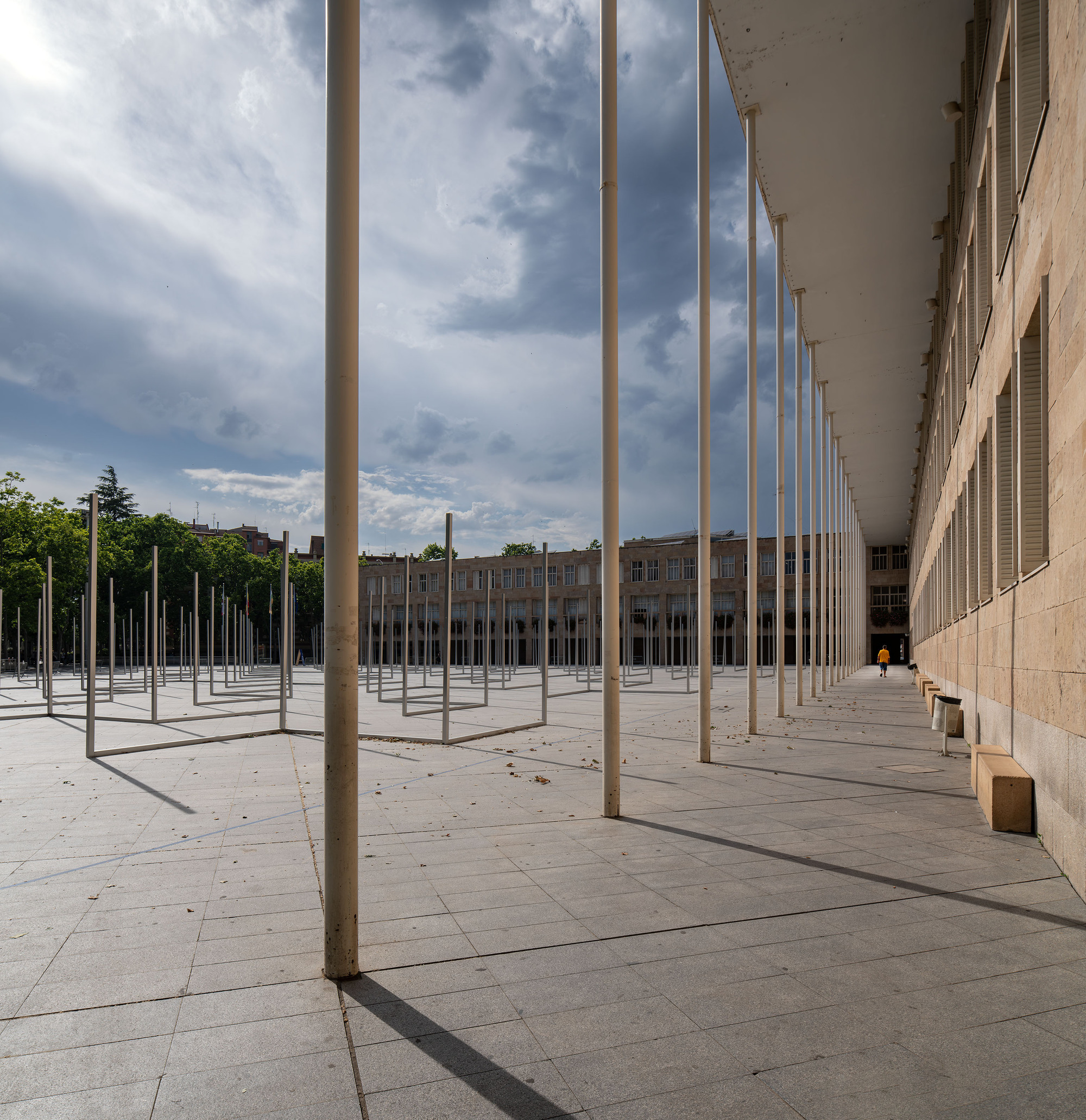 Colonnade of white columns along a stone building edge, with a lone walker in yellow and storm clouds over the plaza