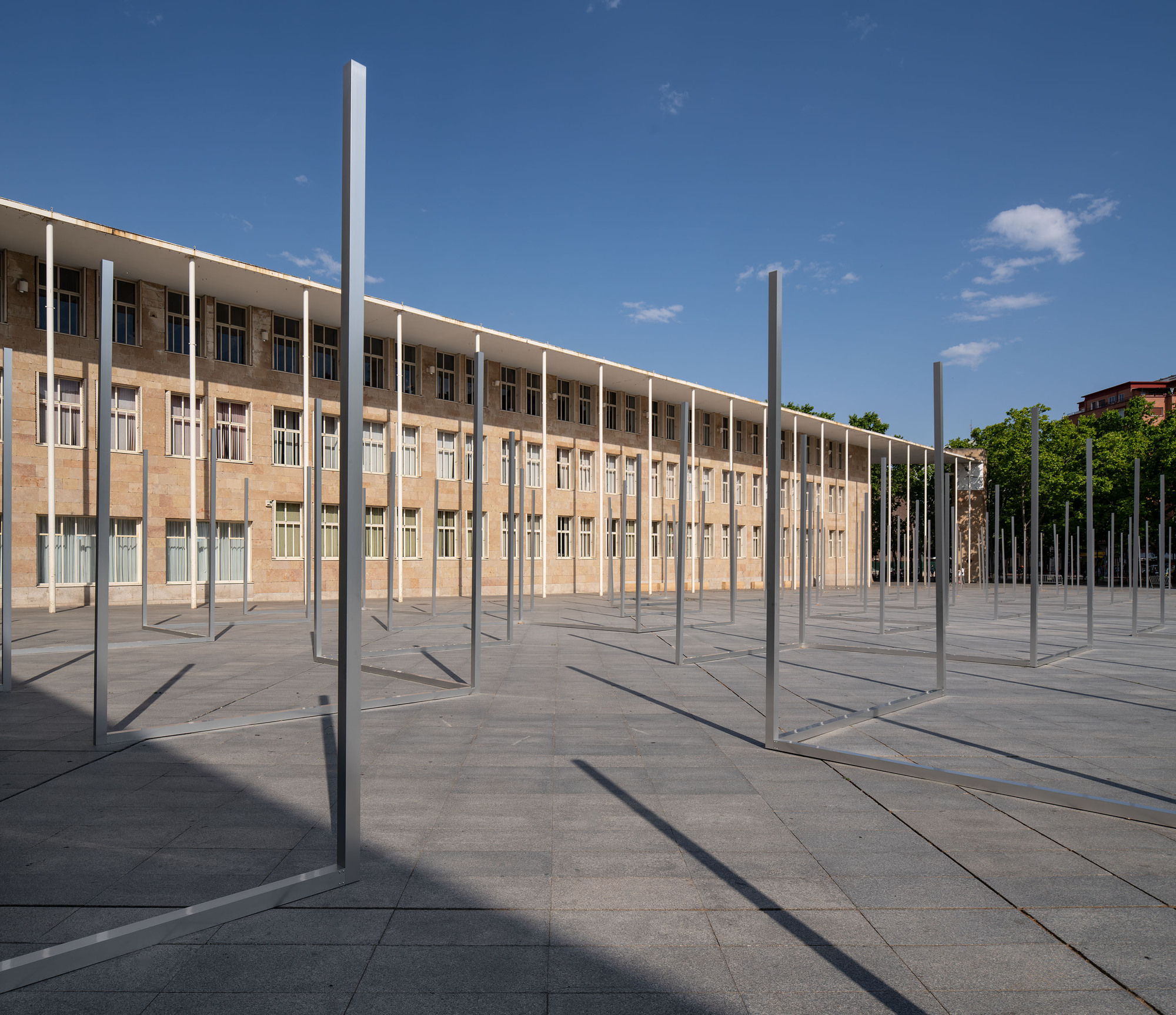 Tall open metal frames rising from the paving, set against a long stone facade and a deep blue sky with scattered clouds
