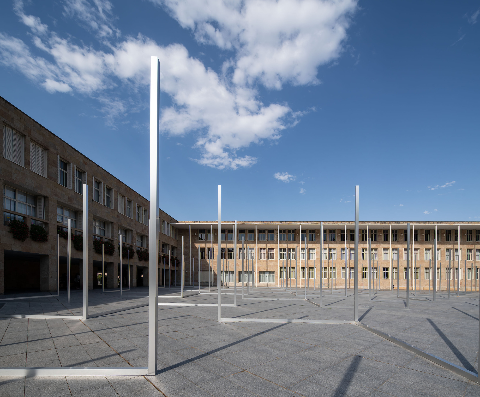 Wide view of slim metal frames scattered across a granite plaza enclosed by stone buildings, beneath a large cloud in blue sky