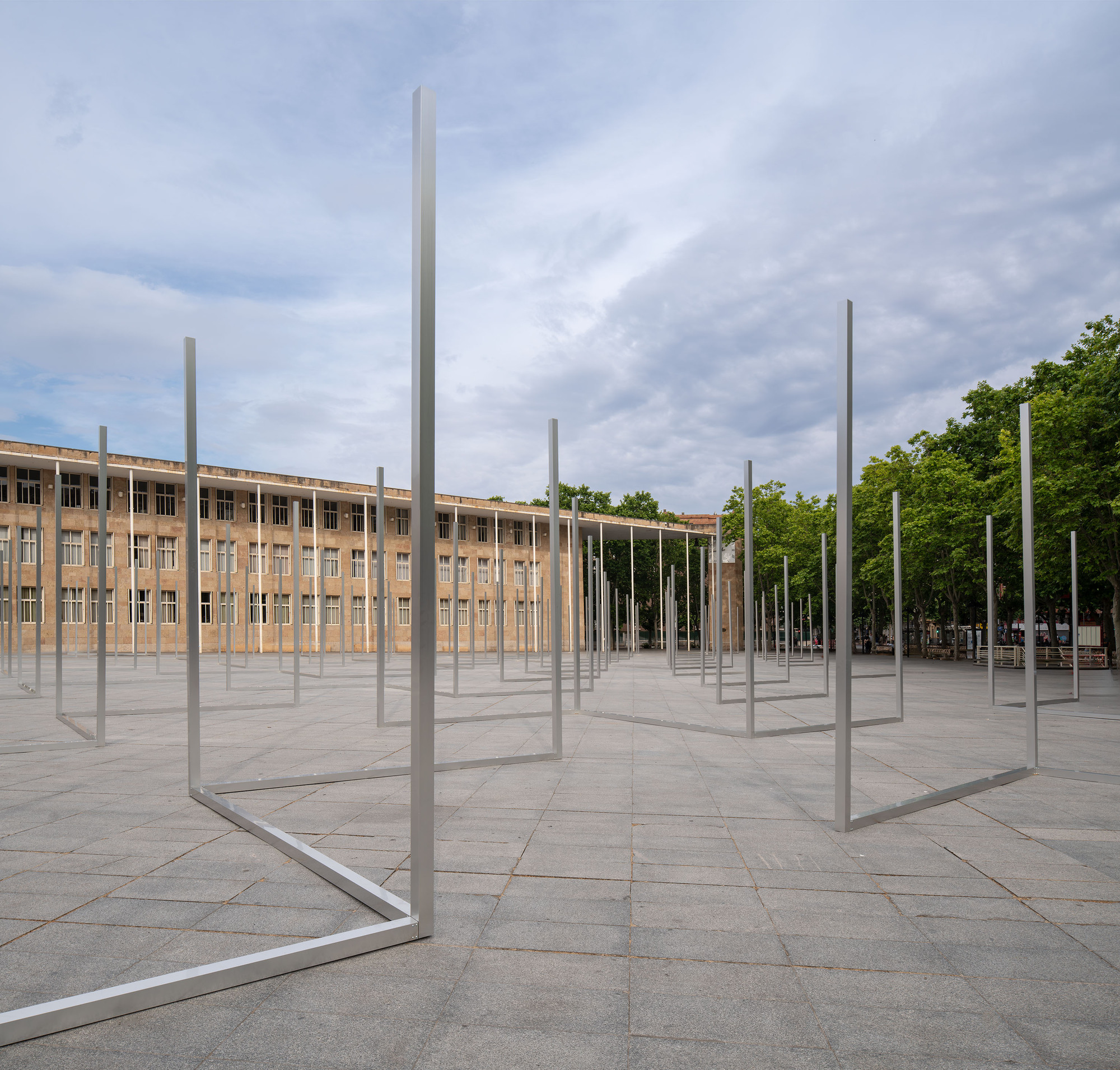 Open steel frames standing on the paved plaza in front of a stone-clad colonnaded building under a cloudy sky