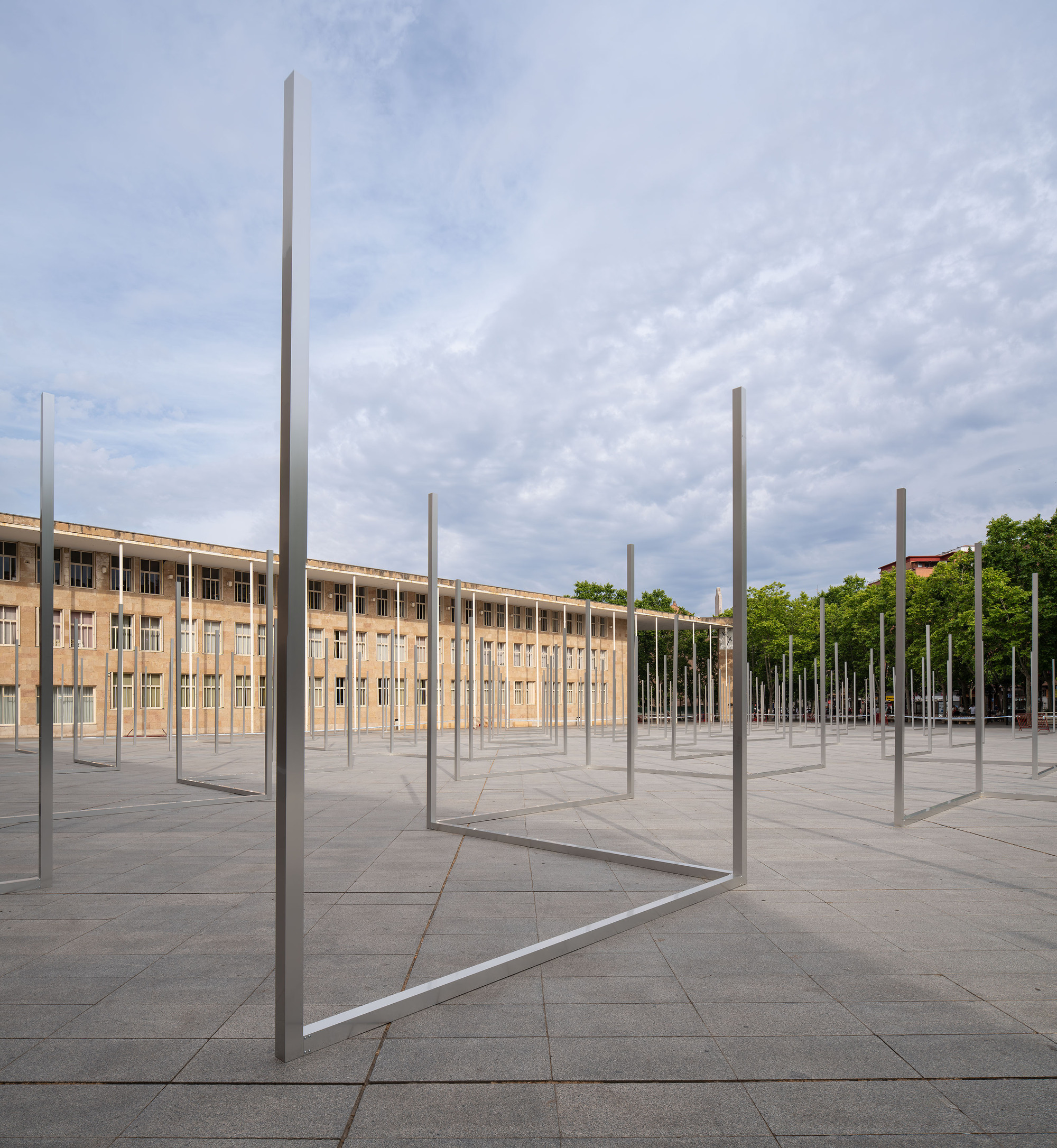 Tall metal L-shaped frames repeating across the paving toward a sandstone building and a row of trees under soft cloud