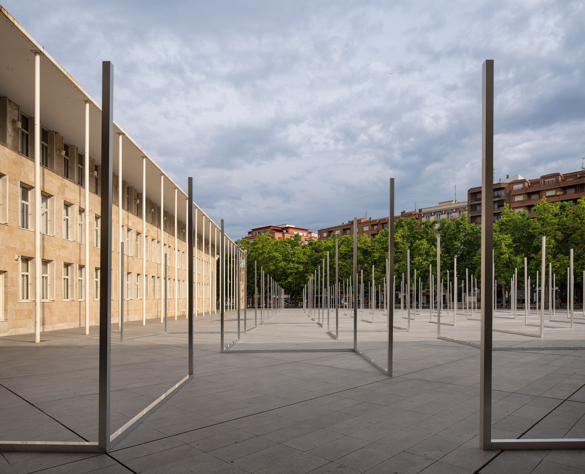 Receding grid of slender steel frames beside a colonnaded stone facade, with green trees and brick apartments beyond in overcast light