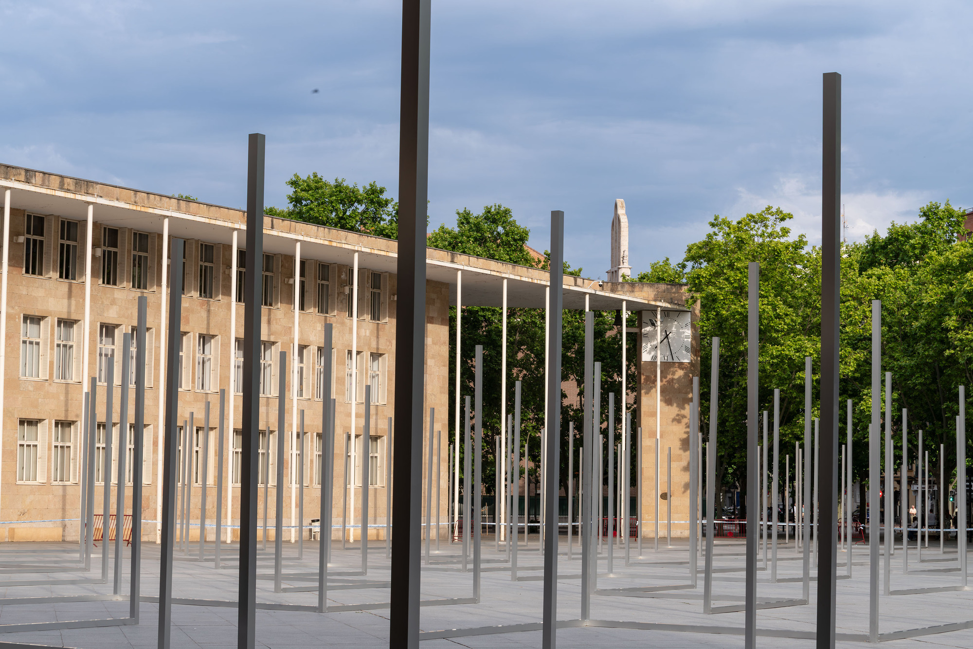 Dense field of vertical steel posts in front of a colonnaded facade with a clock and tower, lit by low sun
