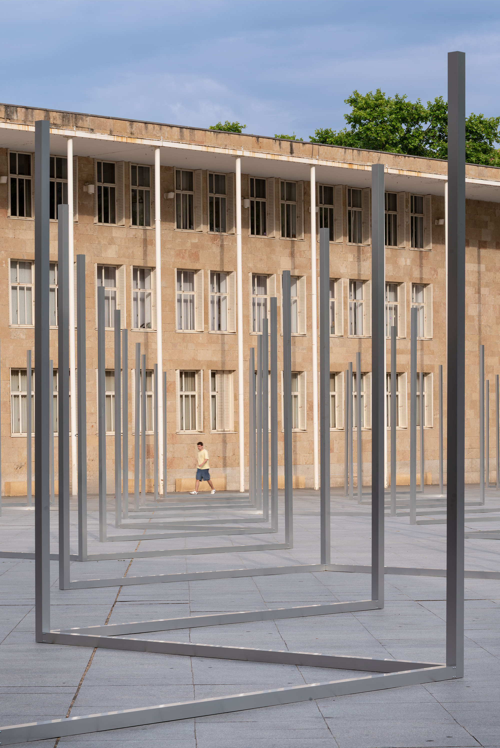 Rows of slender steel frame outlines standing on a paved plaza before a stone-clad building, with a pedestrian walking past in evening light