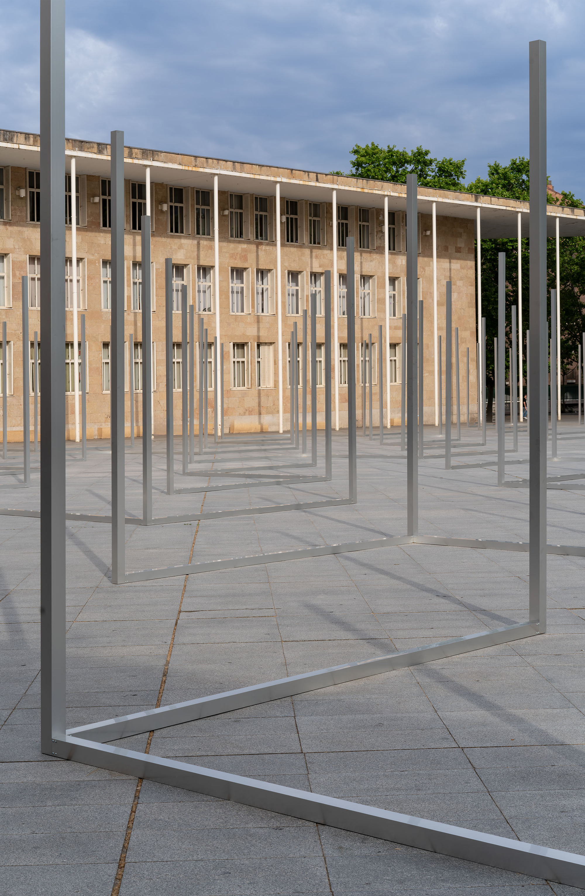 Low view of open steel rectangular frames on granite paving, their long shadows falling toward a colonnaded facade under cloudy skies